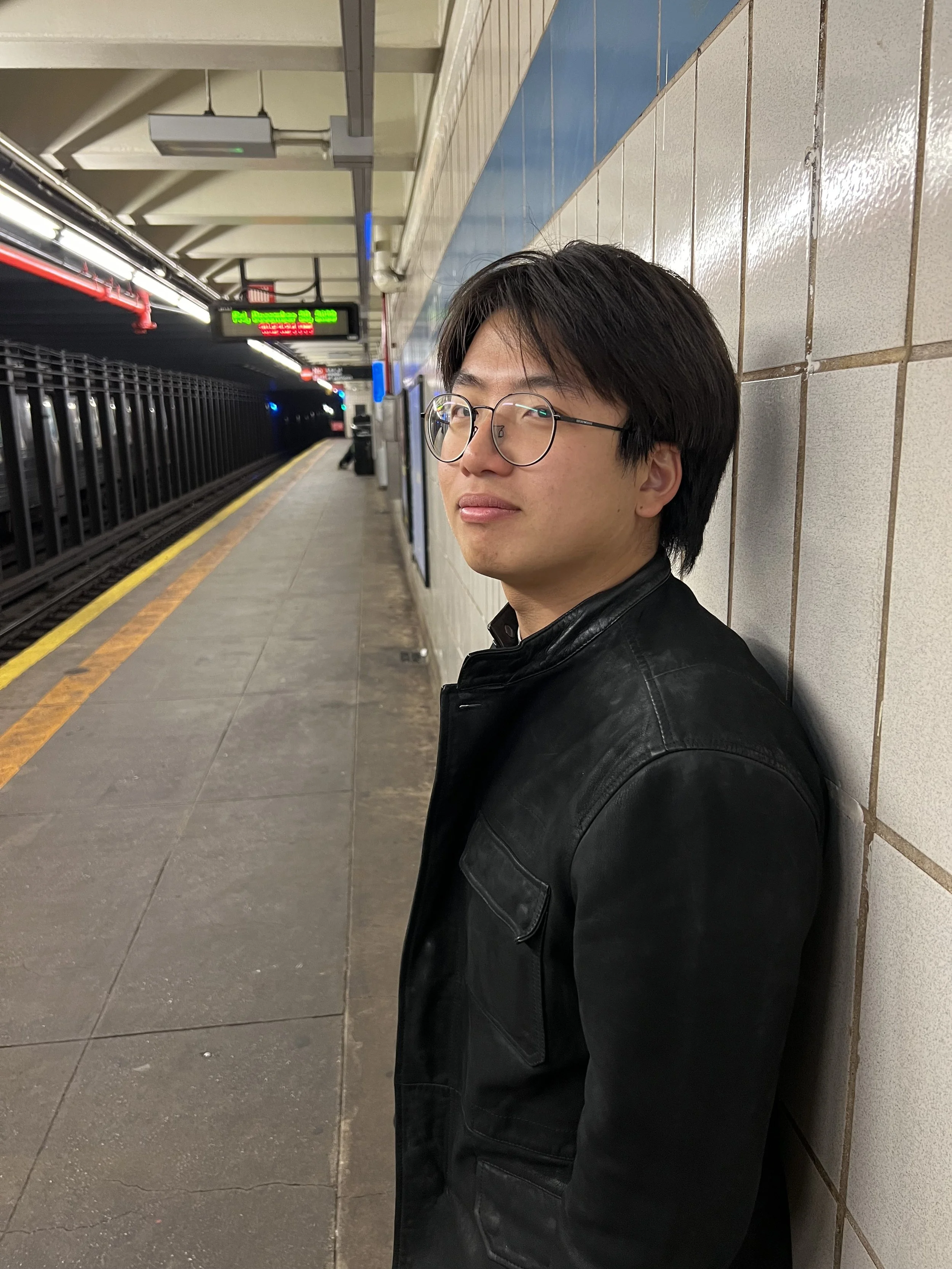 A young man with glasses and dark hair leaning against a tiled wall in a subway station, with a train platform behind him and an electronic sign overhead.