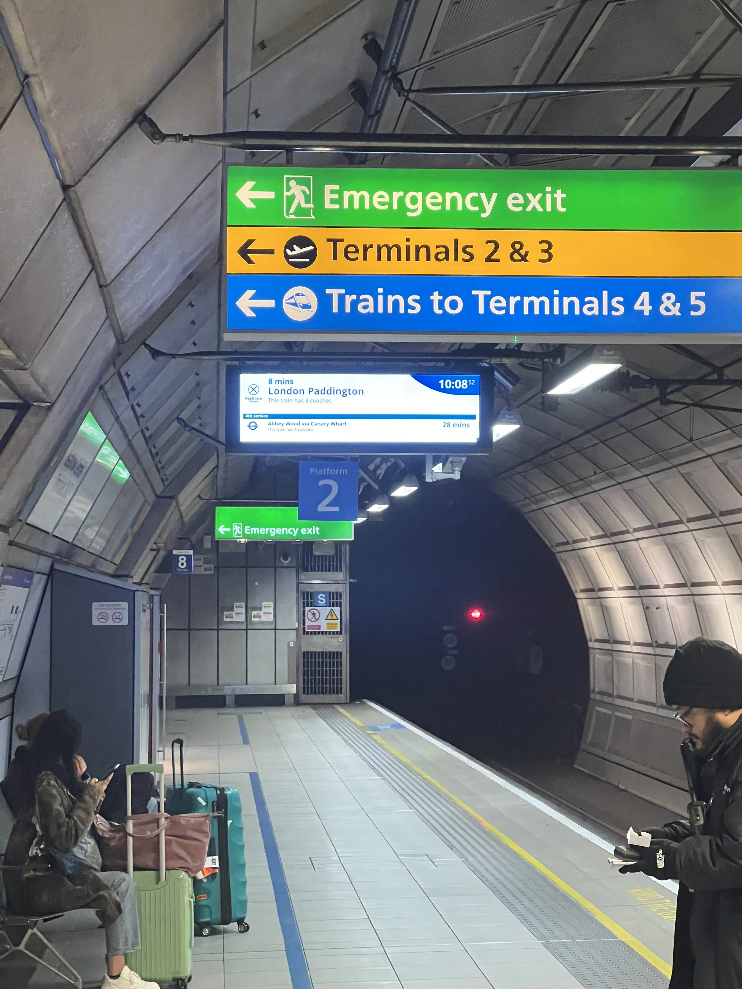 Airport terminal with seating area occupied by travelers, luggage, and digital flight information screens, along with directional signs for emergency exit, terminals 2 & 3, and trains to terminals 4 & 5.