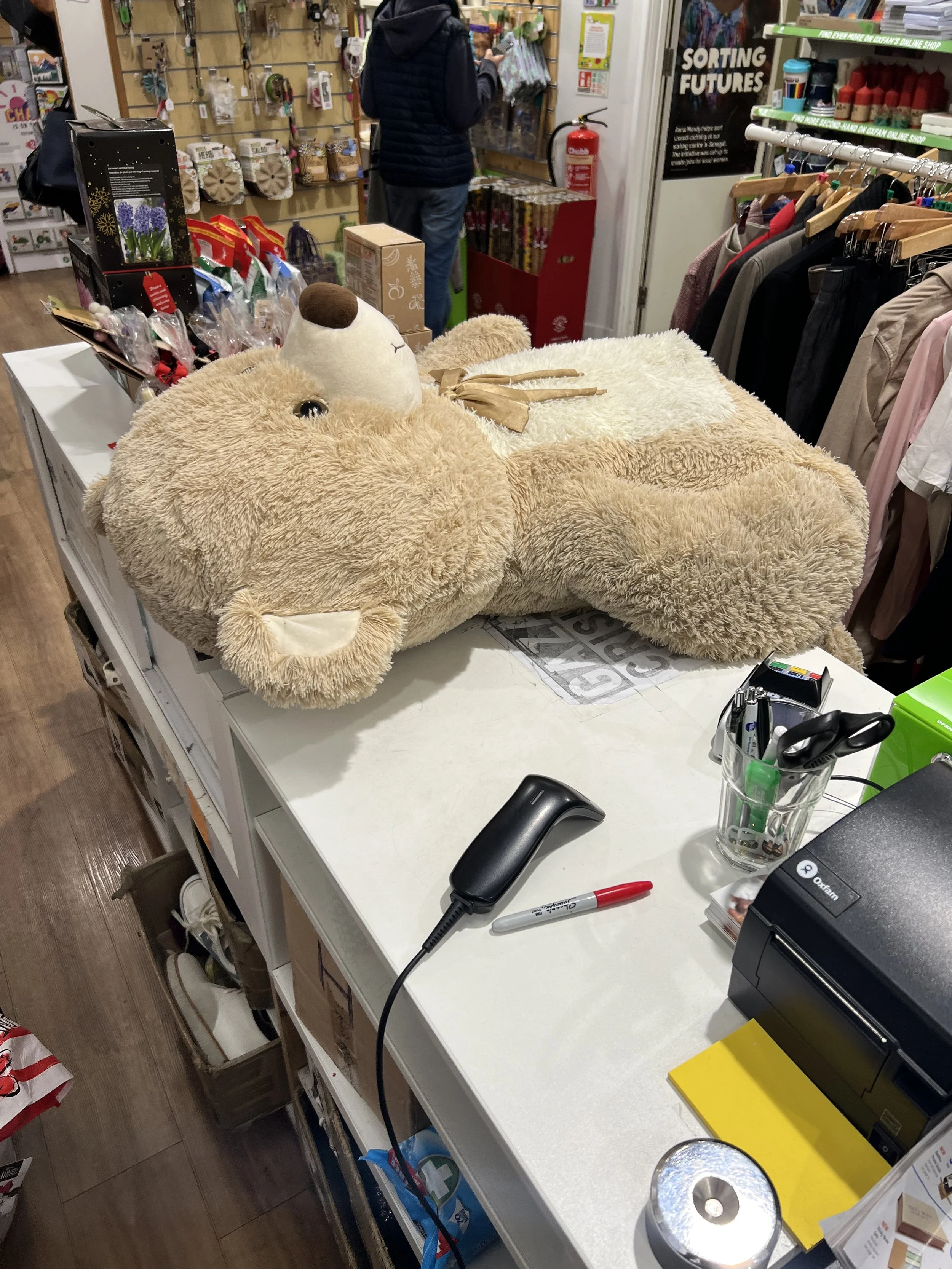 A store checkout counter with a large beige plush bear lying on its back on the counter, surrounded by various items including a cordless phone, markers, and office supplies. In the background, a customer is browsing in the store, and shelves are sto