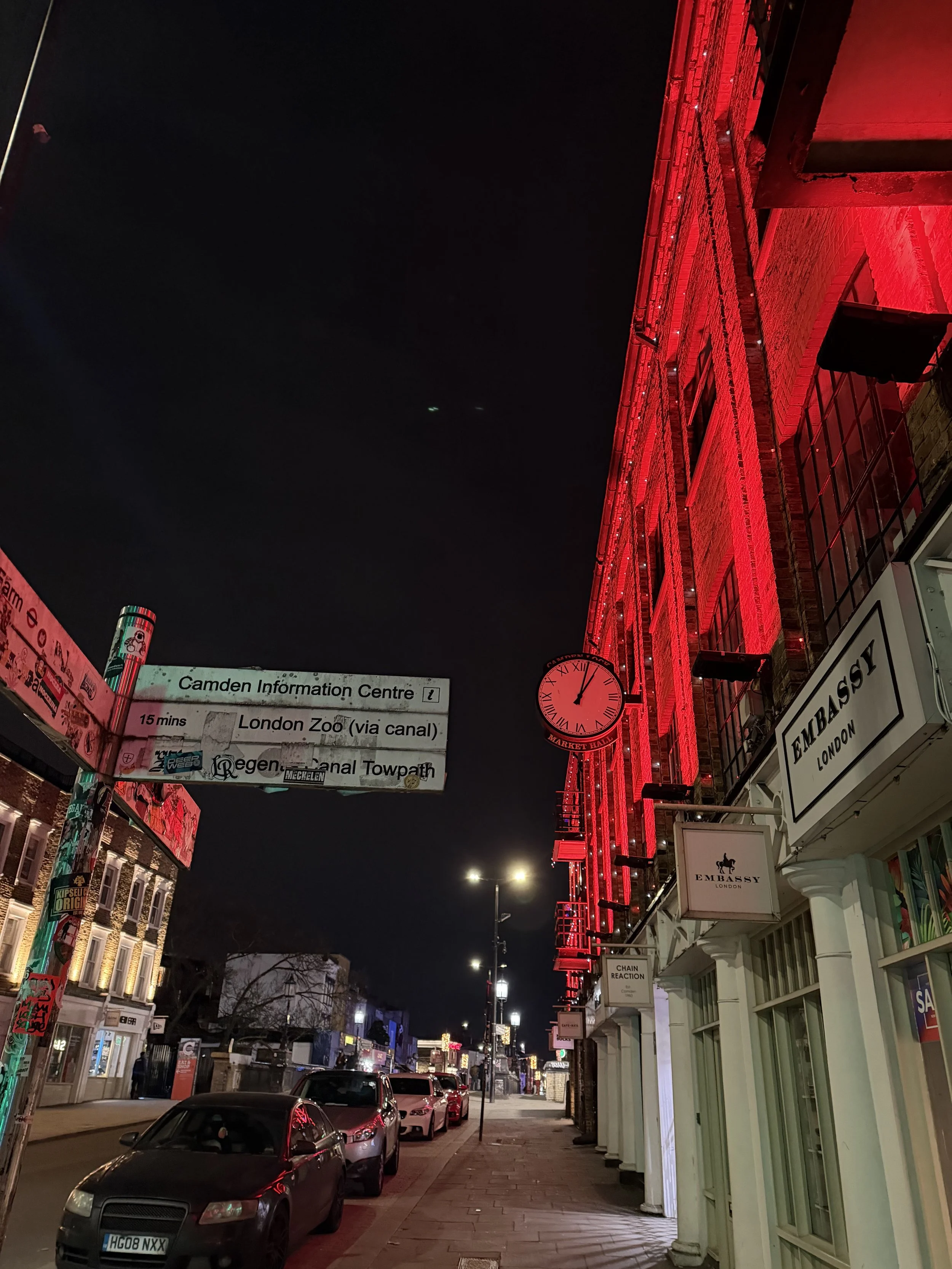 Nighttime street scene in London with illuminated red building, parked cars, street signs, and a clock showing 11:55.