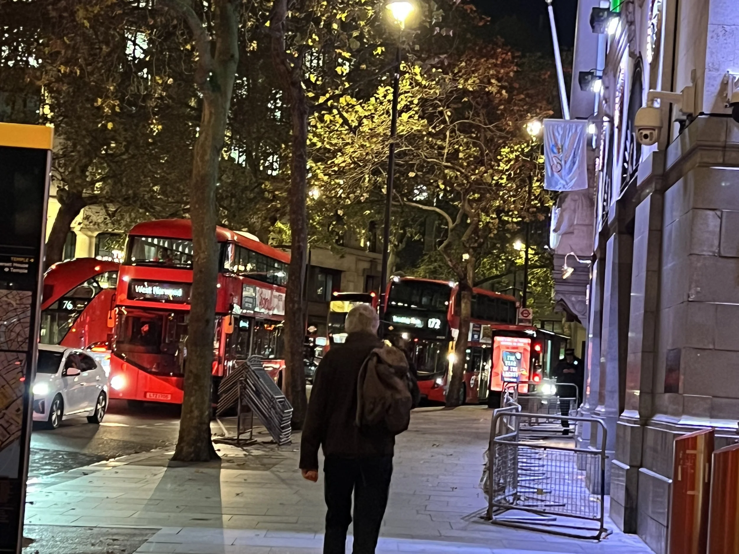Nighttime city street scene with people and vehicles including red double-decker buses, illuminated trees, and streetlights.