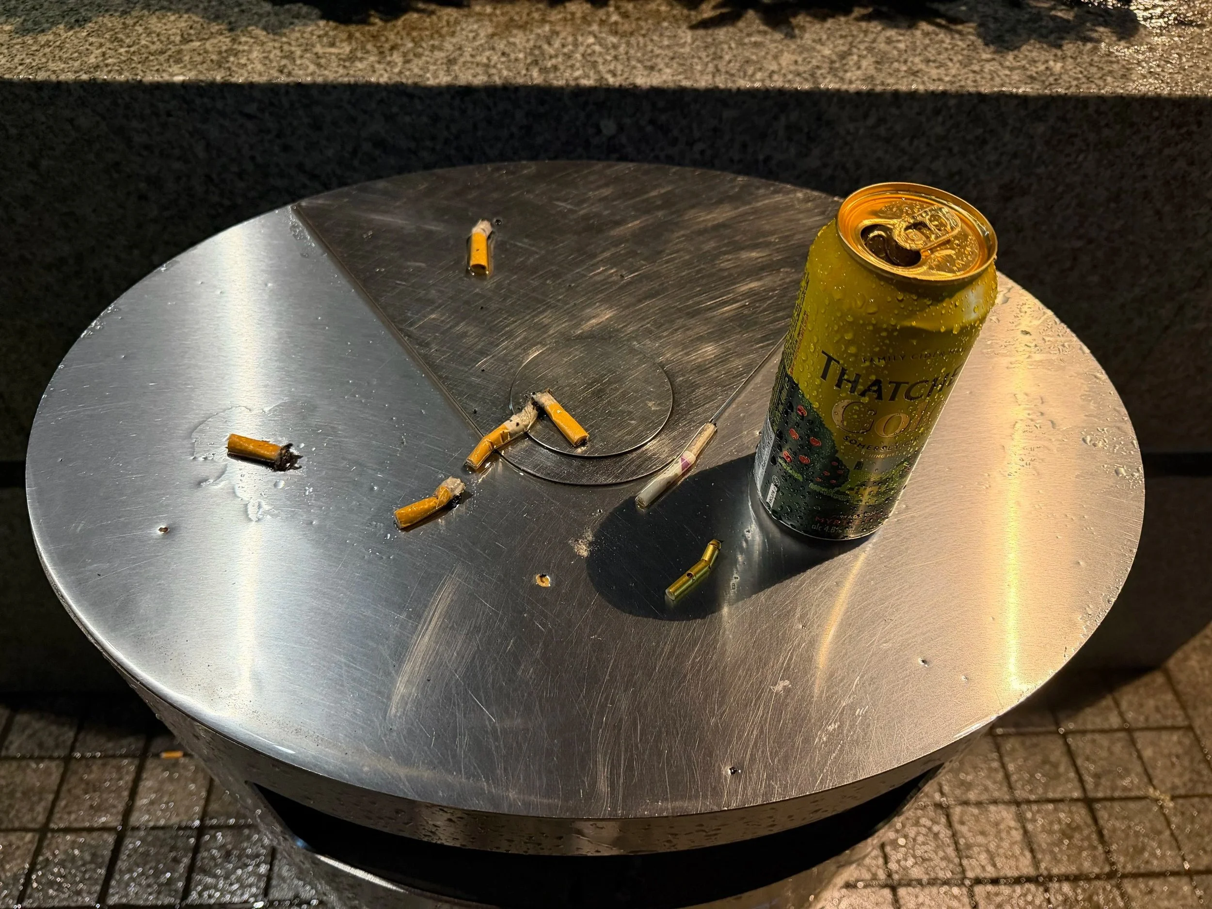 A round metallic table with several cigarette butts and a can of soda on it, situated outdoors.