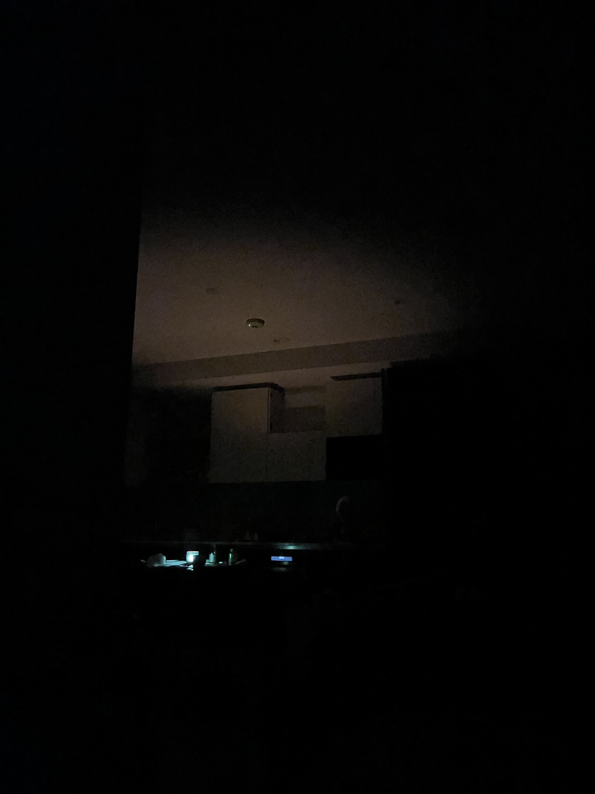 Dimly lit room with faint light from electronic devices on a surface in lower part of the image, ceiling with smoke detector visible, and dark walls and ceiling, with no clear details of objects or people.