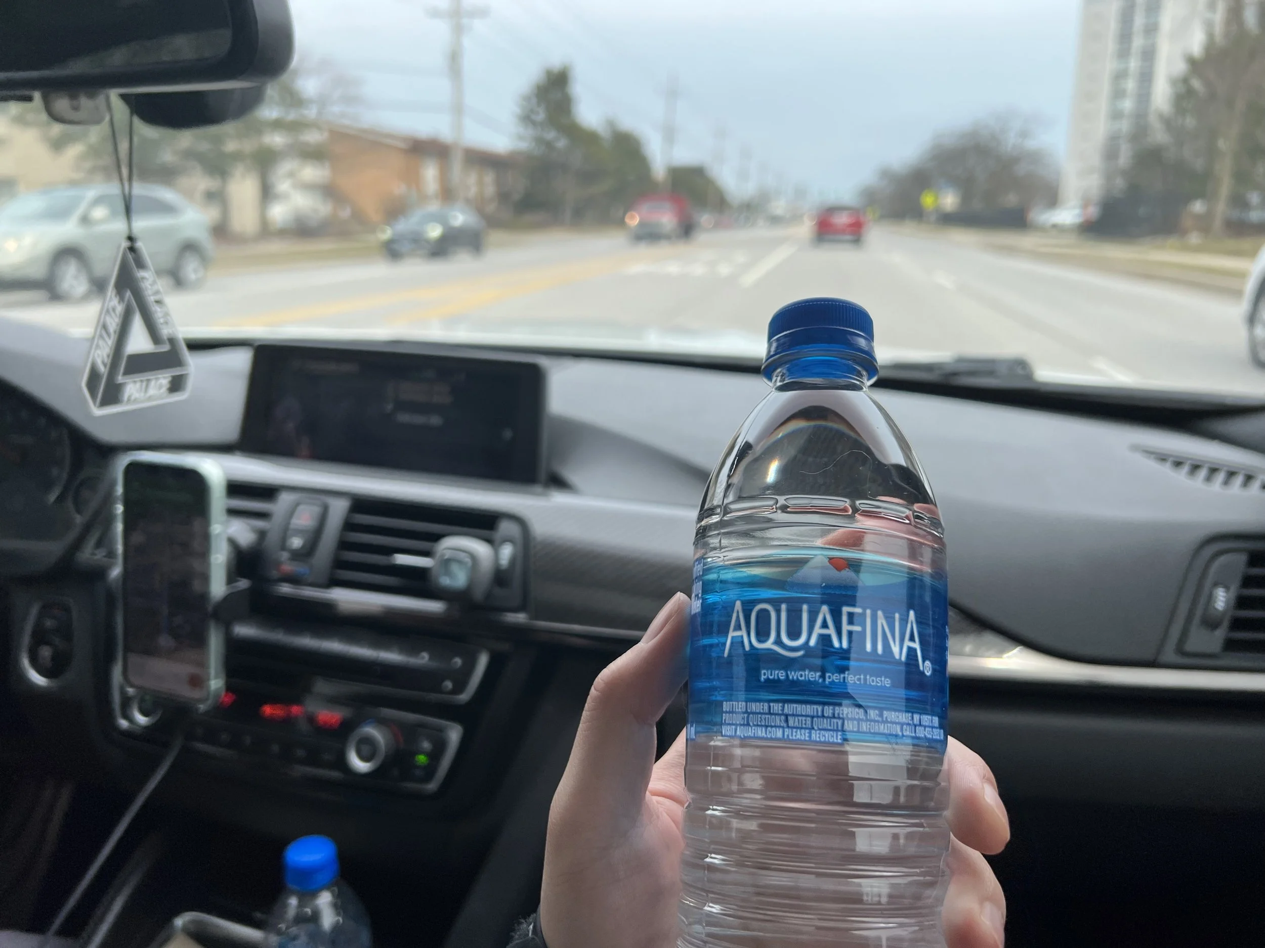 Inside a car, a hand is holding a bottle of Aquafina bottled water with the city road visible through the windshield, some cars and trees in the background.