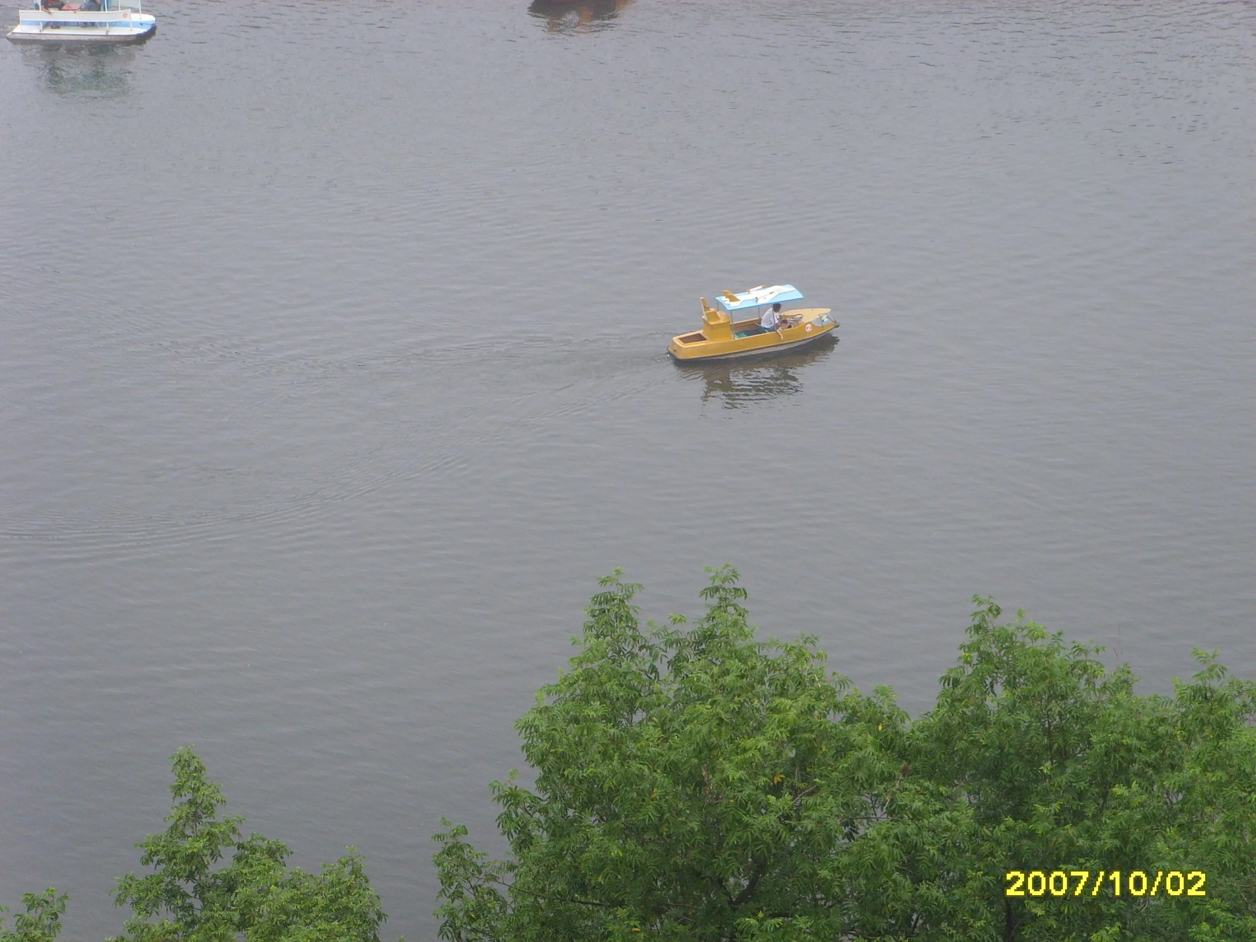 A small yellow boat with a blue roof on a body of water near green trees, with another boat visible in the distance.