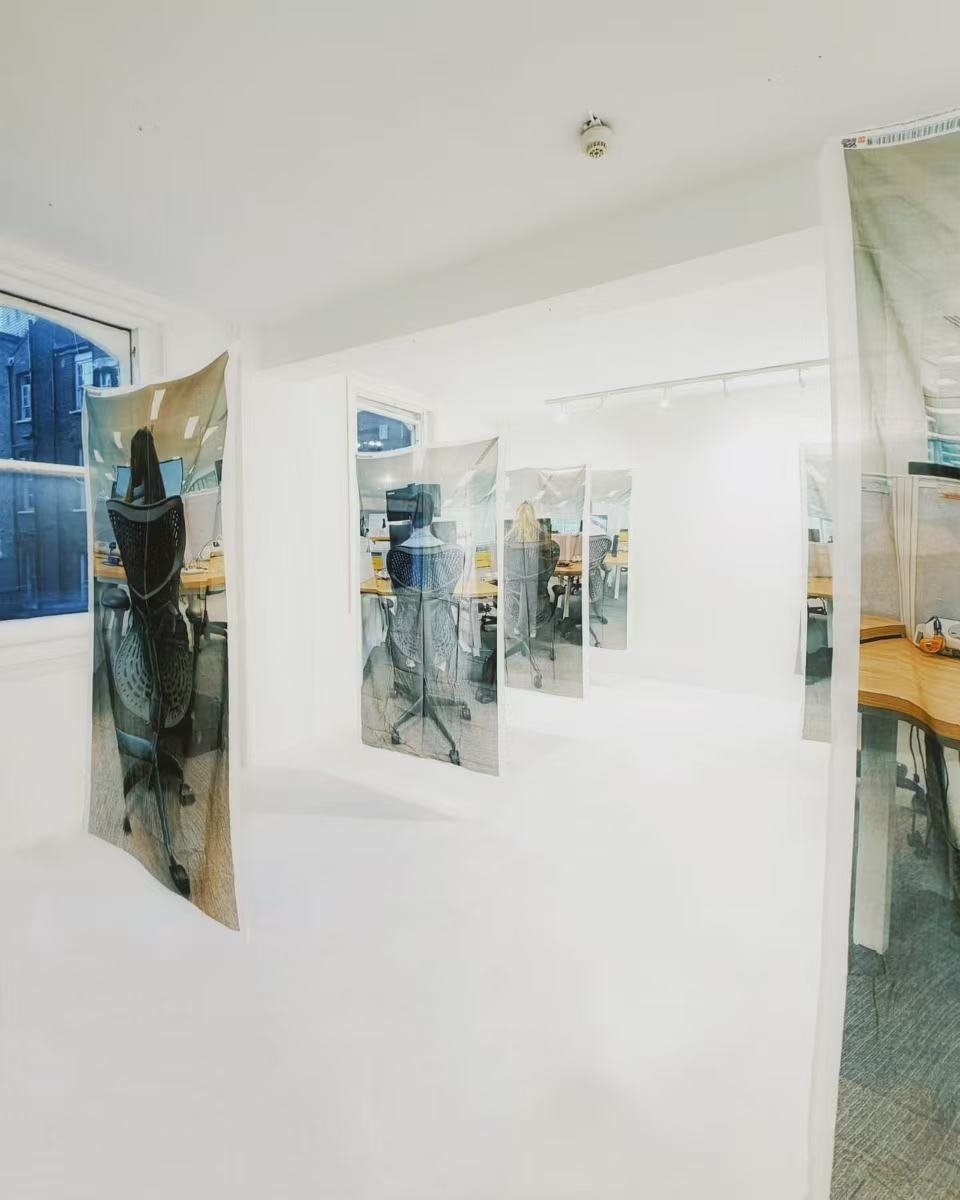 Office with glass panels hanging from ceiling, white walls, windows showing buildings outside, and desks with computers inside.