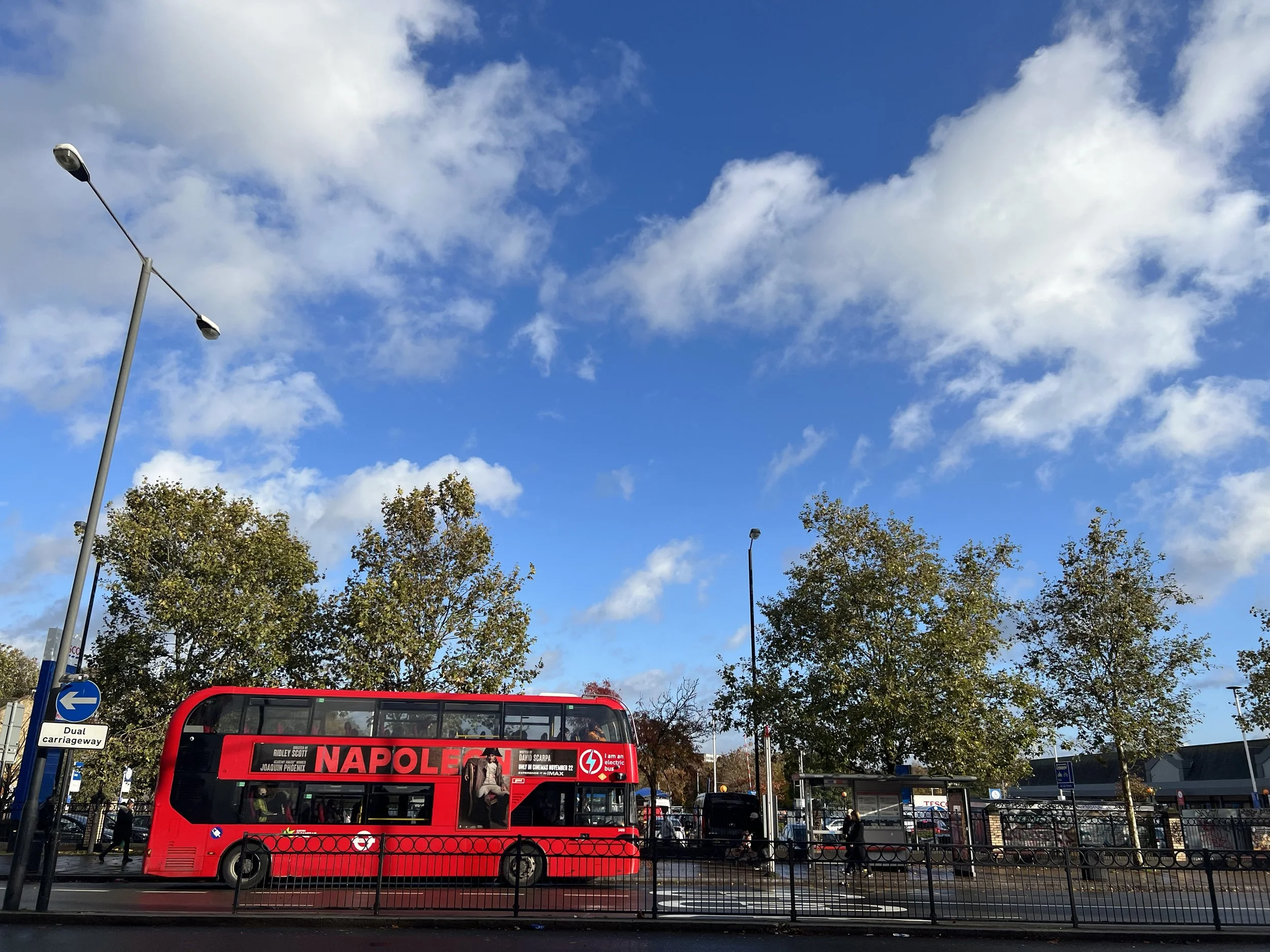 A red double-decker bus with the advertisement for the movie 'Napoleon' in a busy urban setting, with streetlights, trees, and a blue sky with clouds.