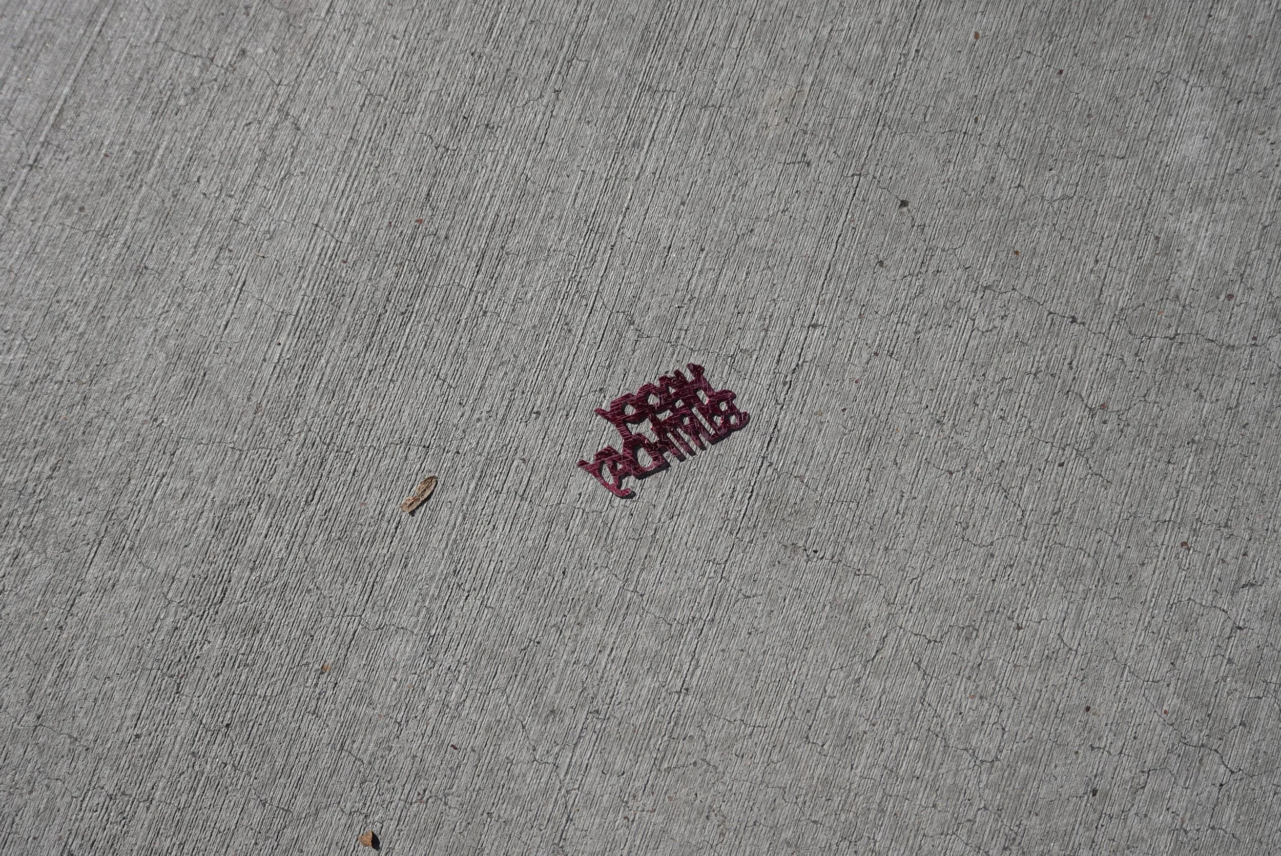 Small red object on concrete sidewalk with partial word visible, possibly part of a sign or message.