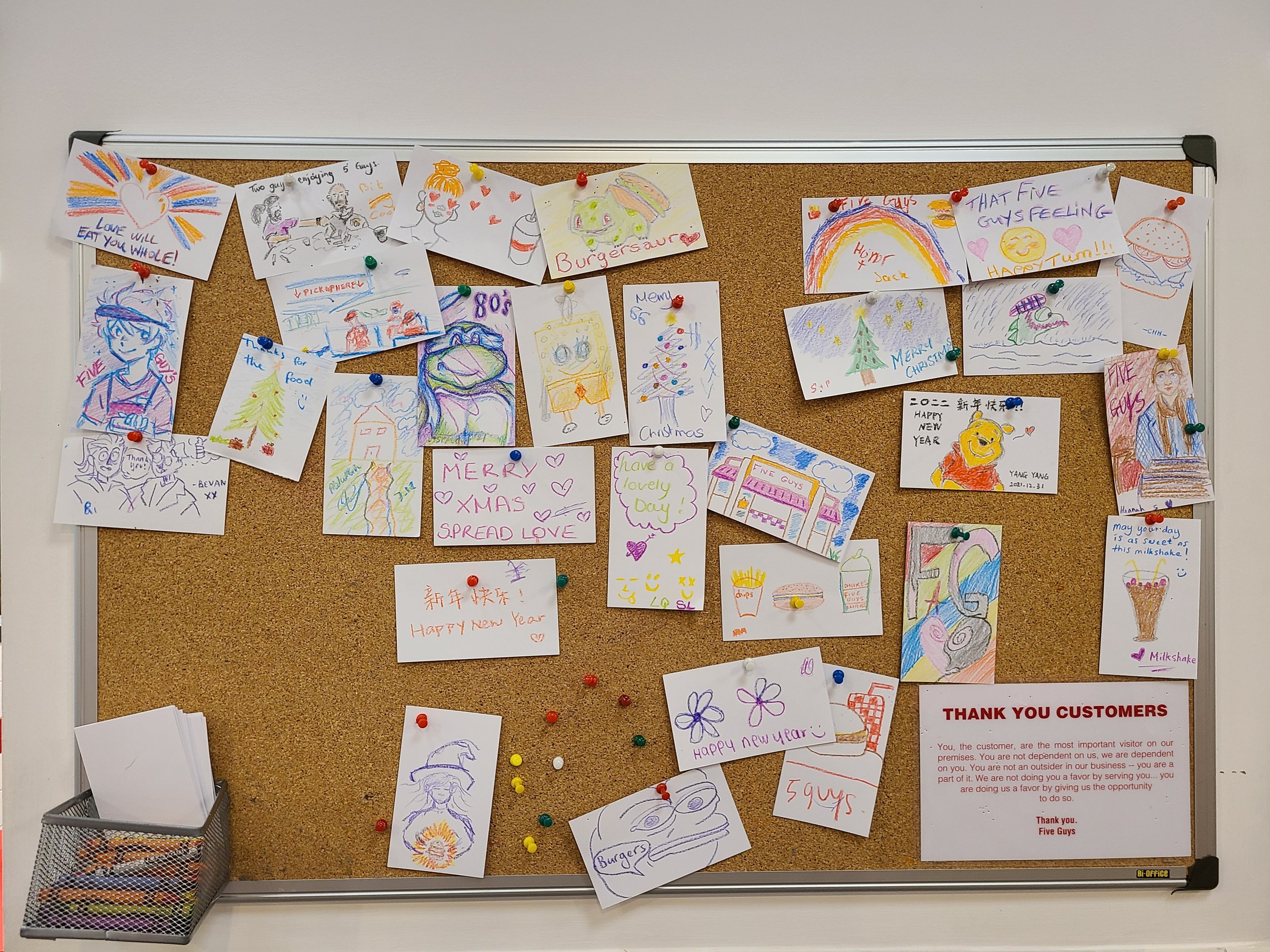 Colorful drawings and notes pinned to a corkboard, including holiday greetings, cartoon characters, and various doodles.