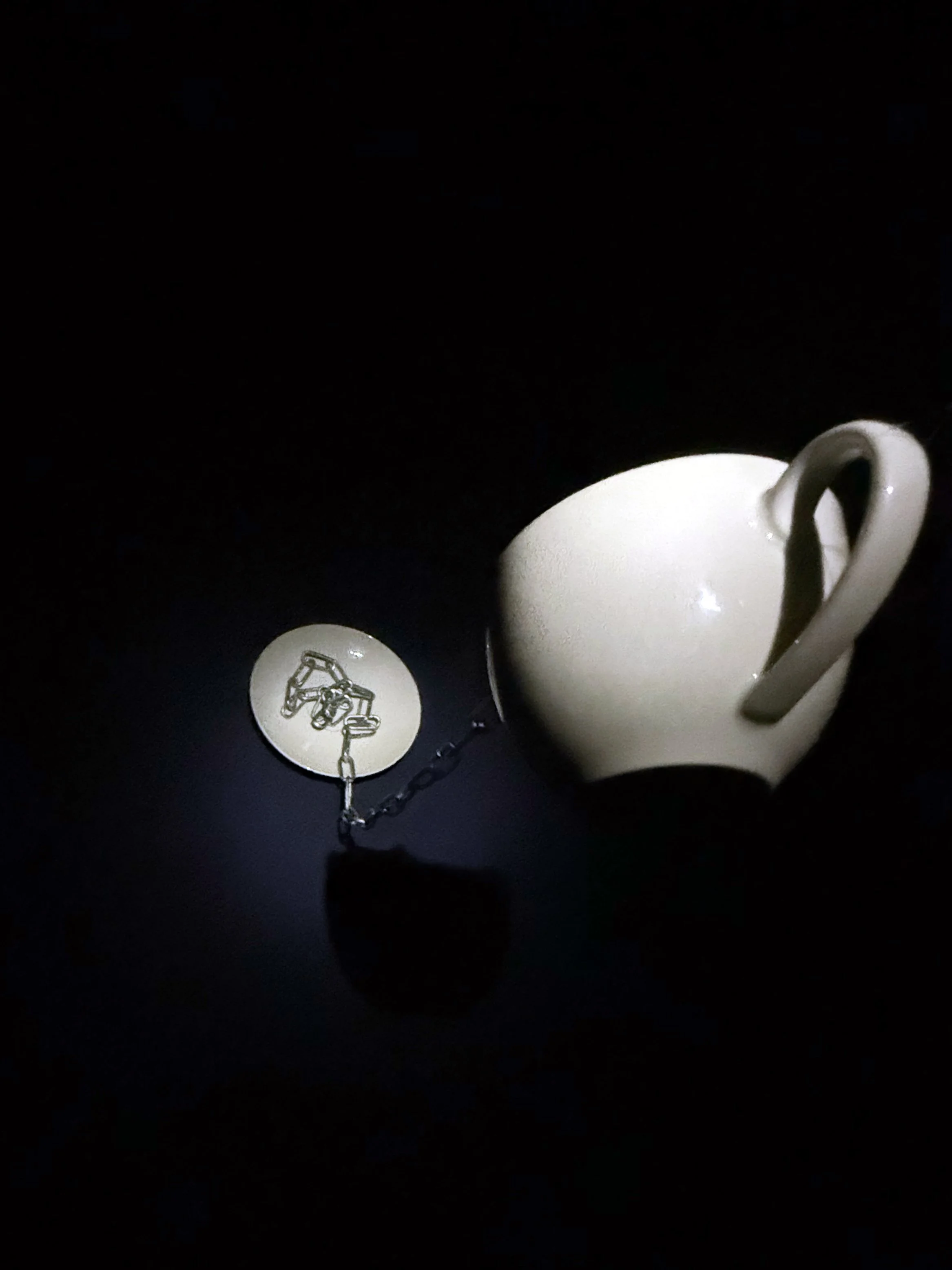 A white coffee mug with a handle, hanging from a chain tied to a metal ring in a dark environment, illuminated by a small light source.