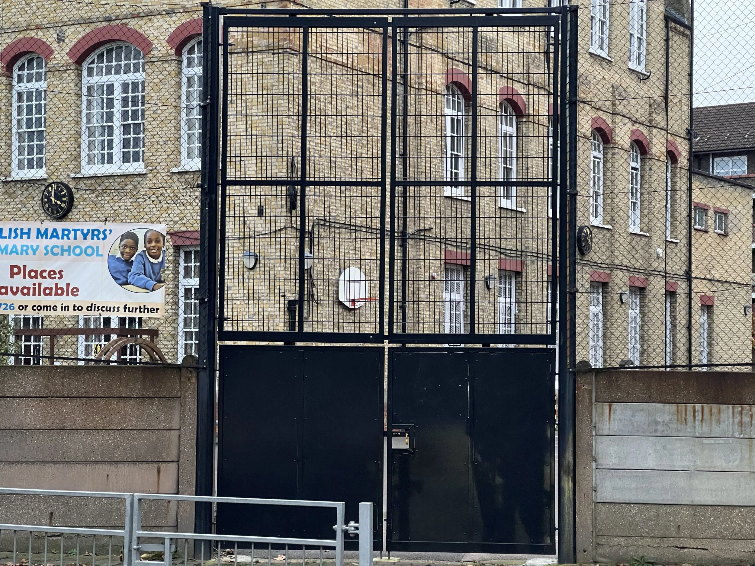 A black metal gate in front of a brick building with white windows and red accents, a clock on the left side, and a sign featuring children in school uniforms.