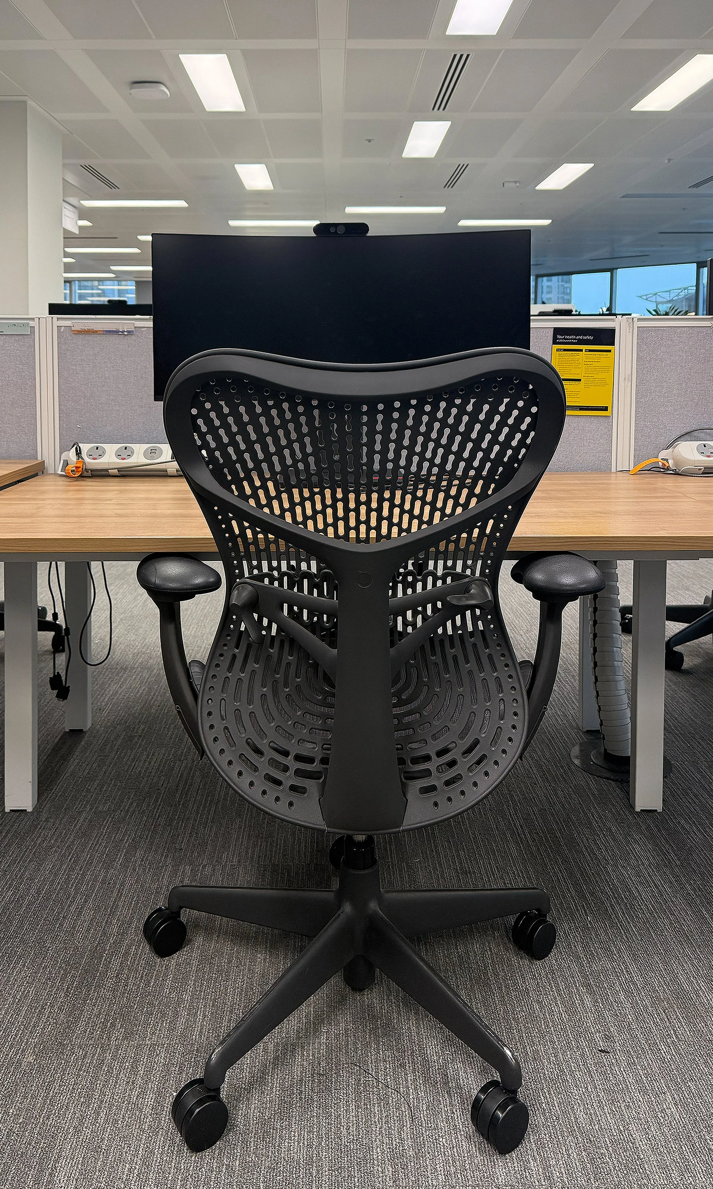 An empty office cubicle with a black ergonomic office chair facing a desk. The desk has a large computer monitor, multiple power outlets, and some cables. Behind the desk are gray partition panels, with a yellow safety notice on one. The office has a