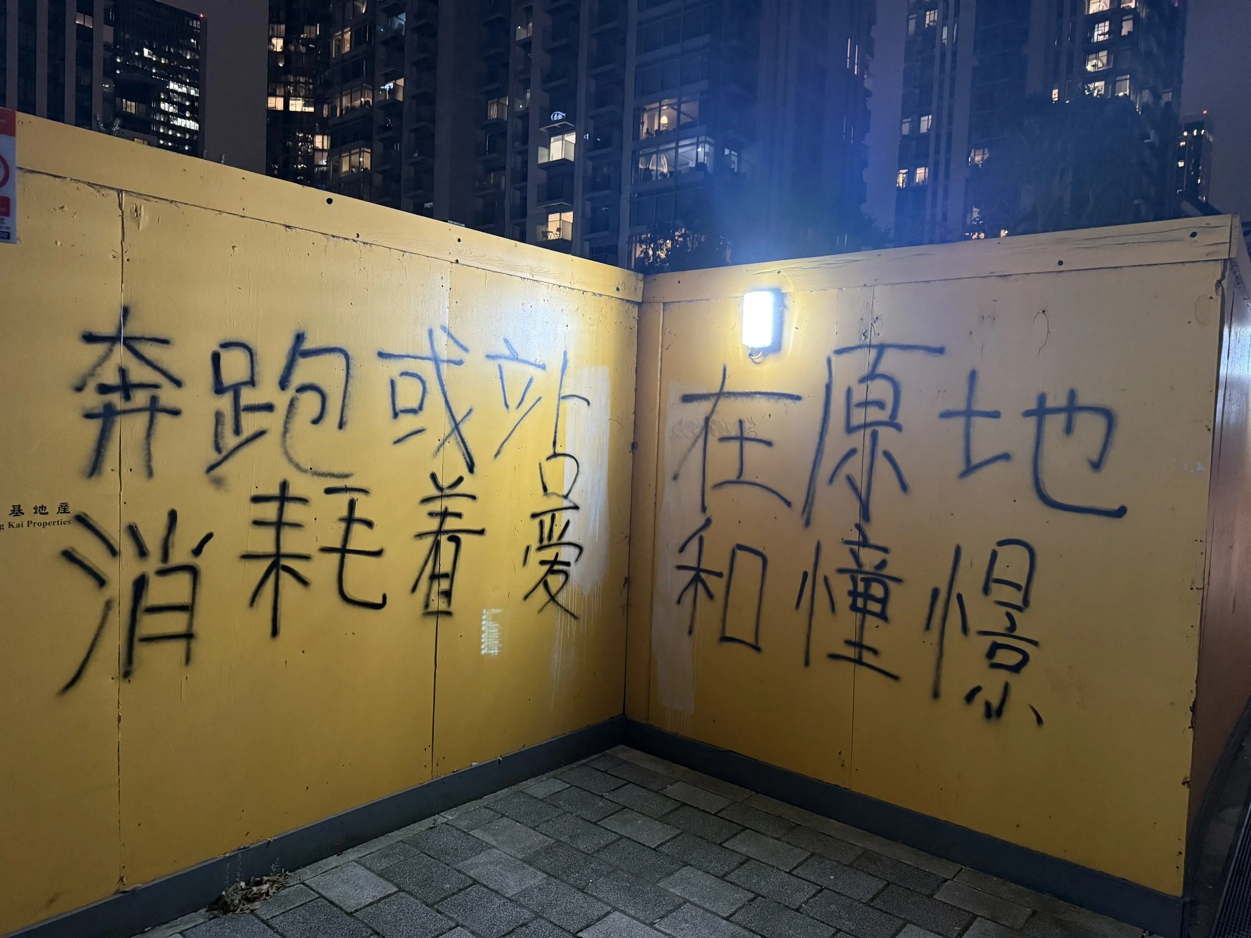 Yellow construction barrier with black graffiti Chinese characters in an urban area at night, with high-rise buildings lit in the background.