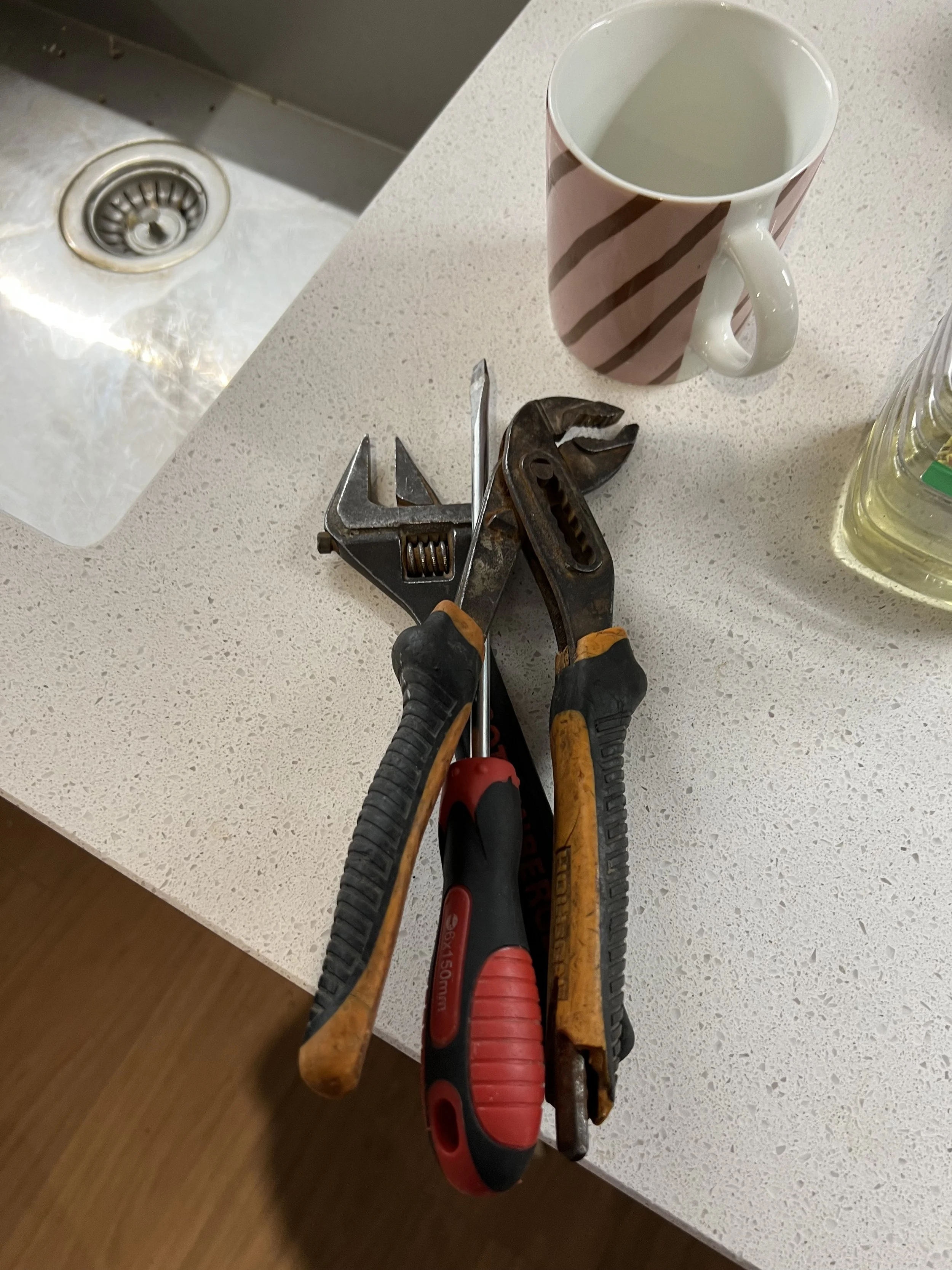 A pair of adjustable wrenches, a screwdriver, and pliers on a white kitchen countertop, with a mug, a bottle of dish soap, and a sink in the background.