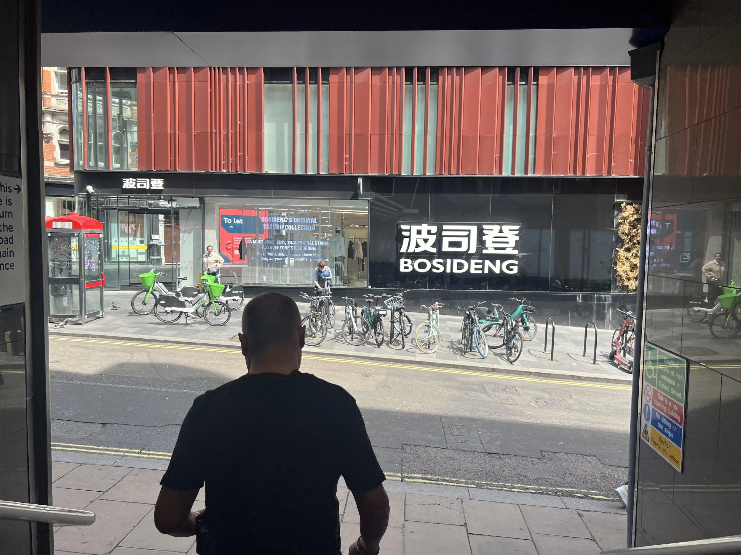Person sitting as viewed from behind outside a building, looking at a busy street scene with bicycles parked in front, a bus stop, and a modern building with prominent red and black exterior design.