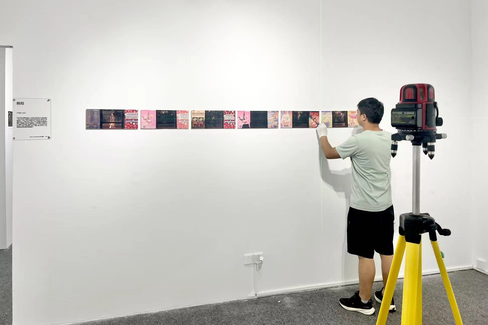 A man standing in an art gallery, looking at a horizontal row of small photographs or artwork on a white wall. He is wearing a white t-shirt, black shorts, and black sneakers. There is a red and black laser level on a tripod to his right and a plaque