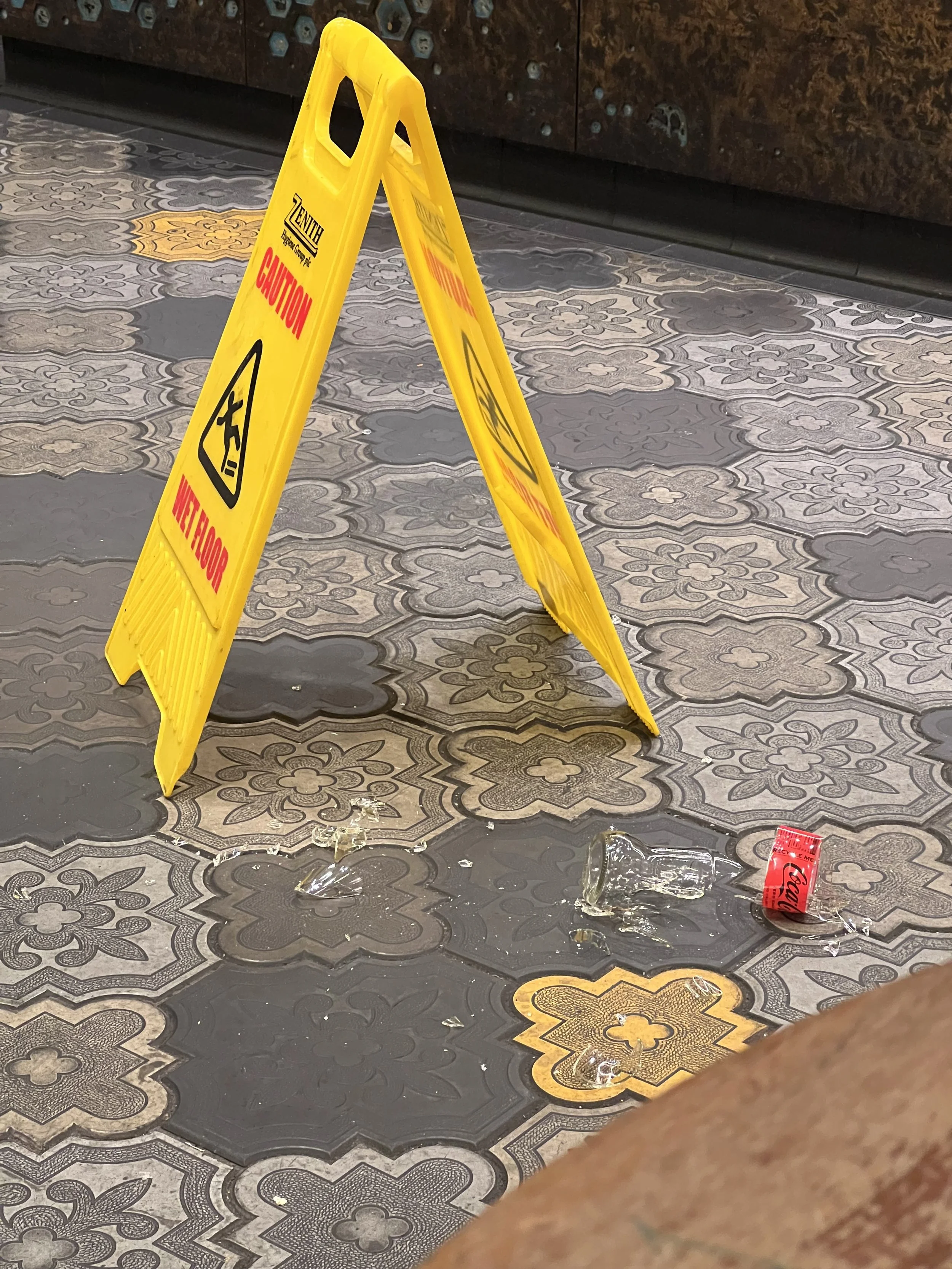 A yellow caution sign on a tile floor with spilled glass and a can, indicating a wet or hazardous surface.