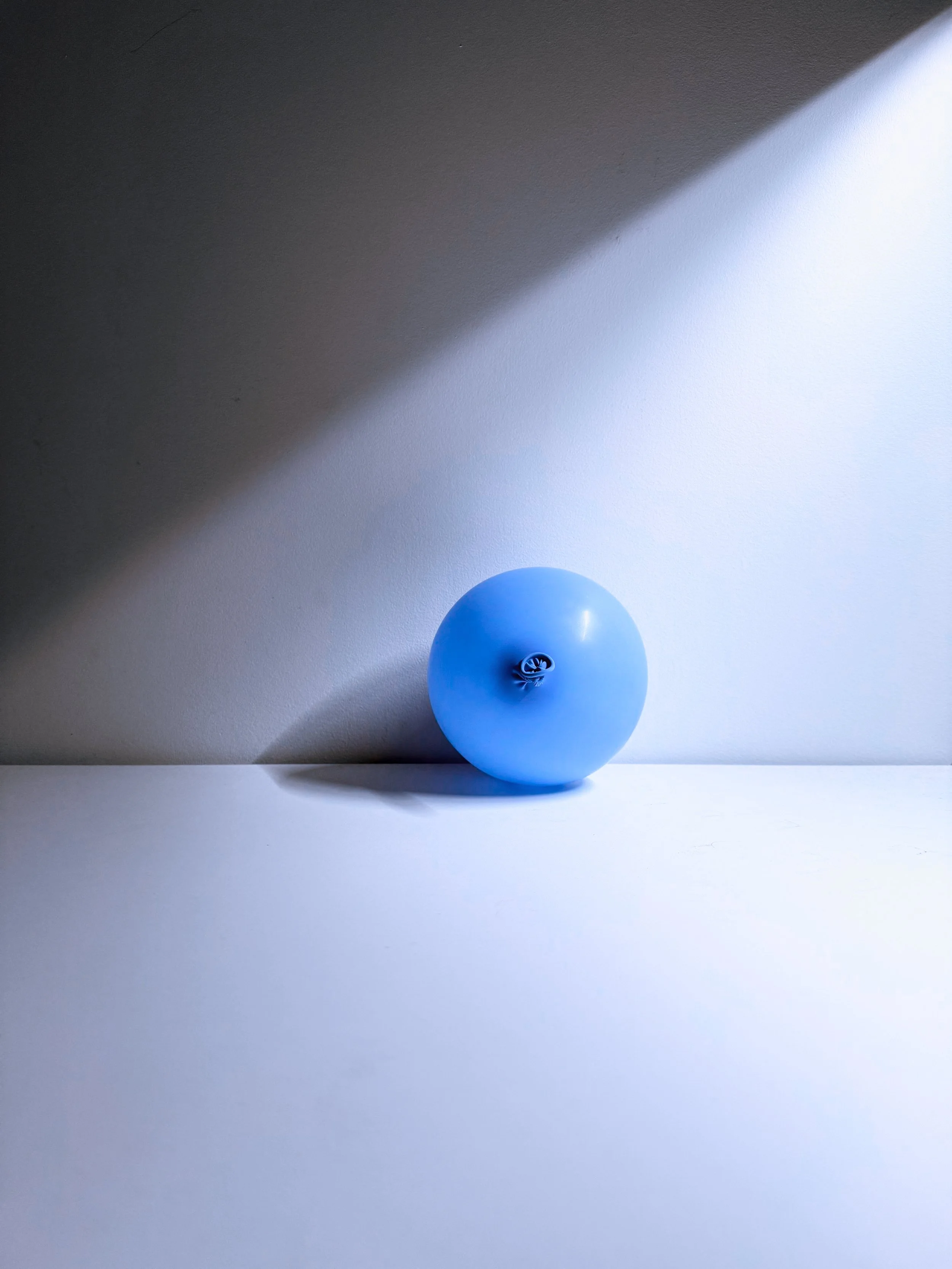 A blue balloon standing on a white surface against a white wall, with a light and dark shadow cast on the wall.