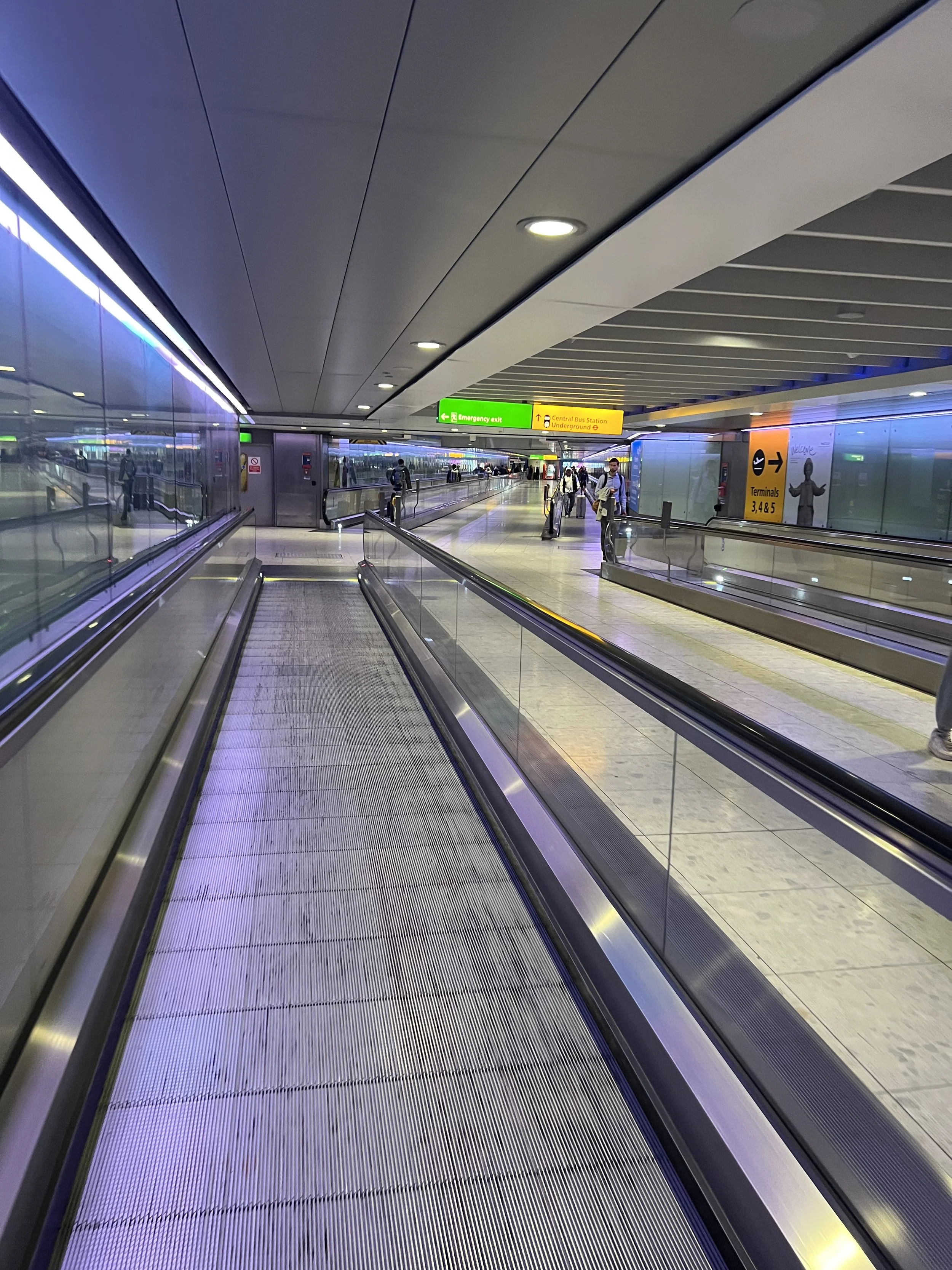 Interior of a modern airport or train station with a moving walkway, passengers waiting and walking, signage for emergency exit and bus or underground train station, glass walls, and ceiling lights.