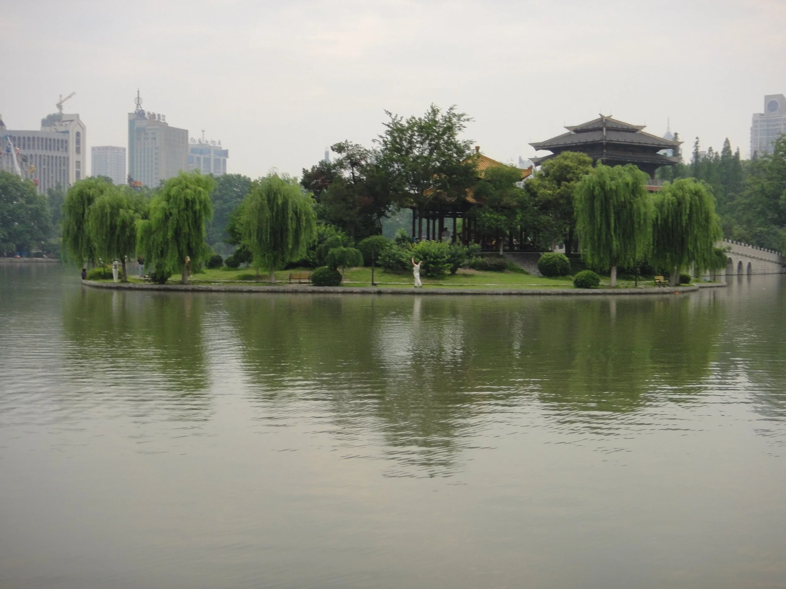 A scenic view of a traditional pagoda-style structure and a pavilion on an island connected by a stone bridge, surrounded by greenery and trees with a city skyline in the background, over a calm body of water.