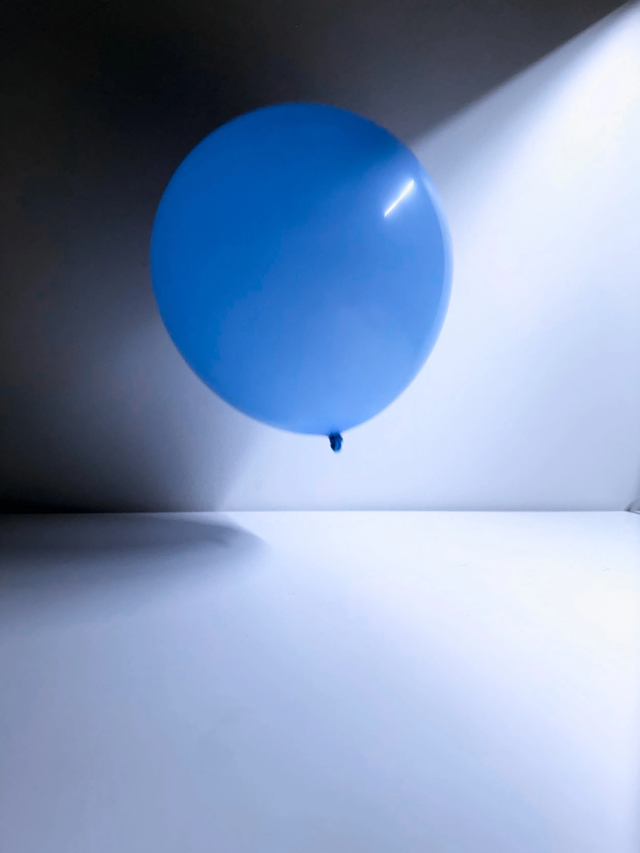 A single blue helium balloon floating in the air with shadow cast on the wall and floor.