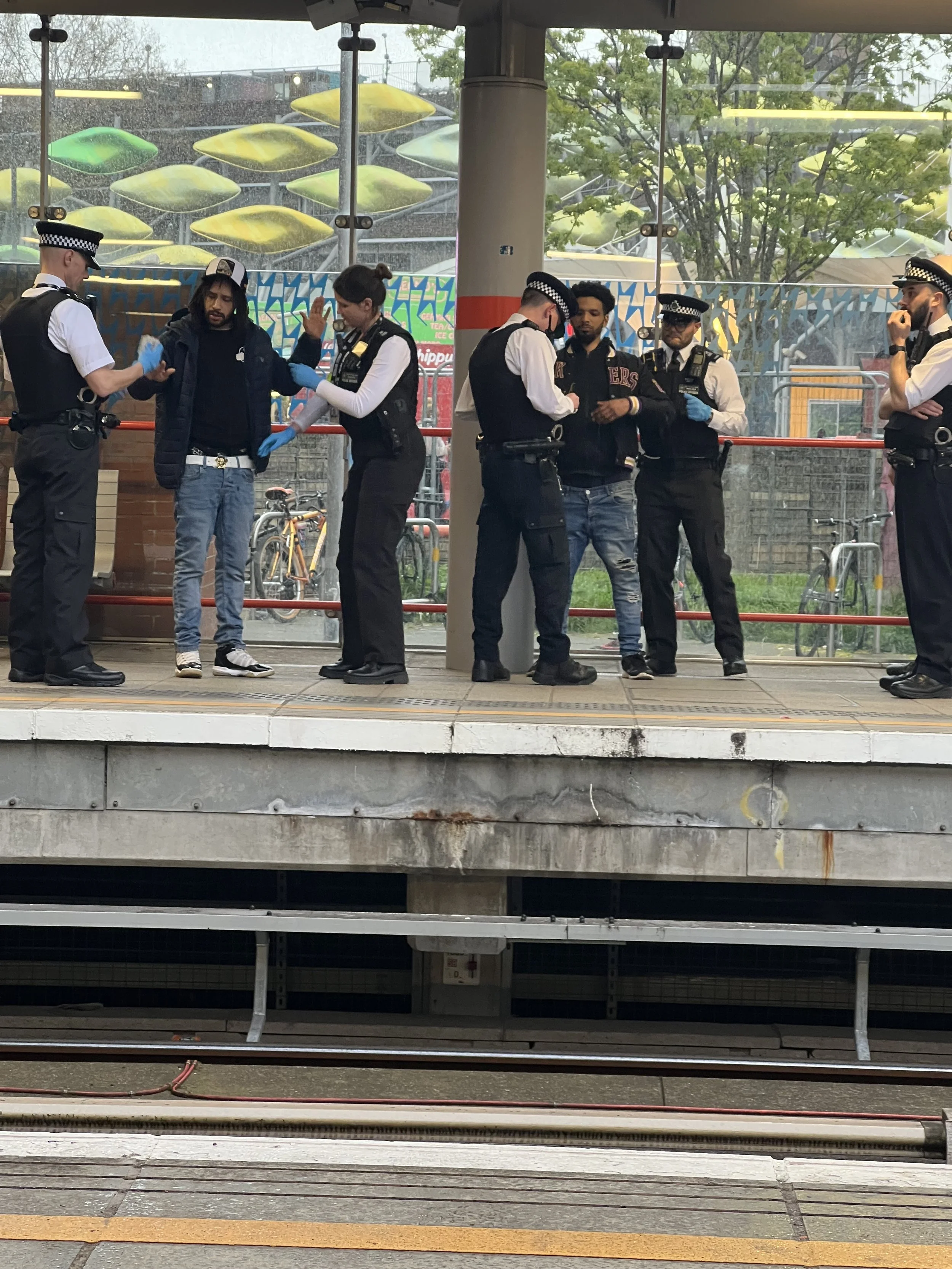 Six police officers and one man are standing on a train platform, with a glass window behind them showing an amusement park ride in the rain.