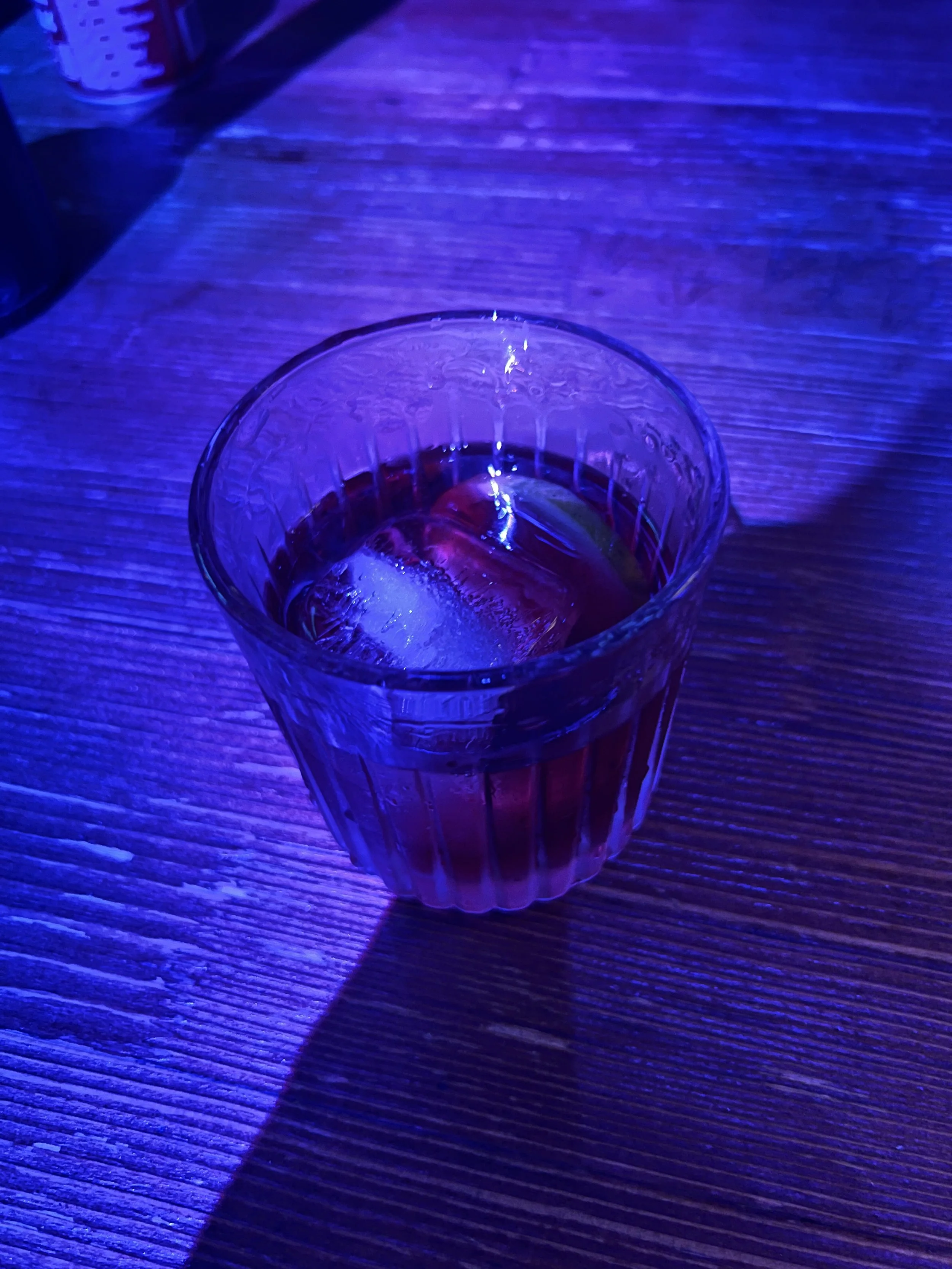 A glass of dark-colored beverage with ice on a wooden table, illuminated with purple and blue lighting.