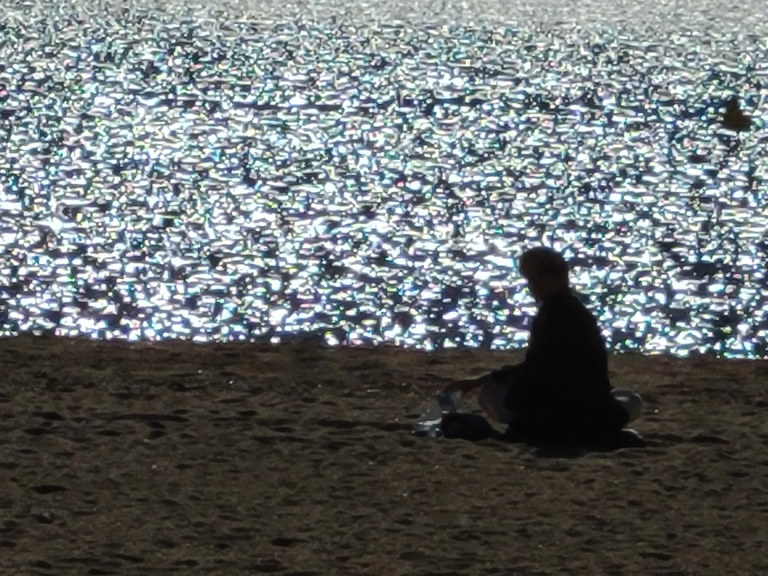 A person sitting on the ground near a large expansive area of reflective, shimmering metallic trash or debris, possibly at a beach or lakeshore during dusk or night.