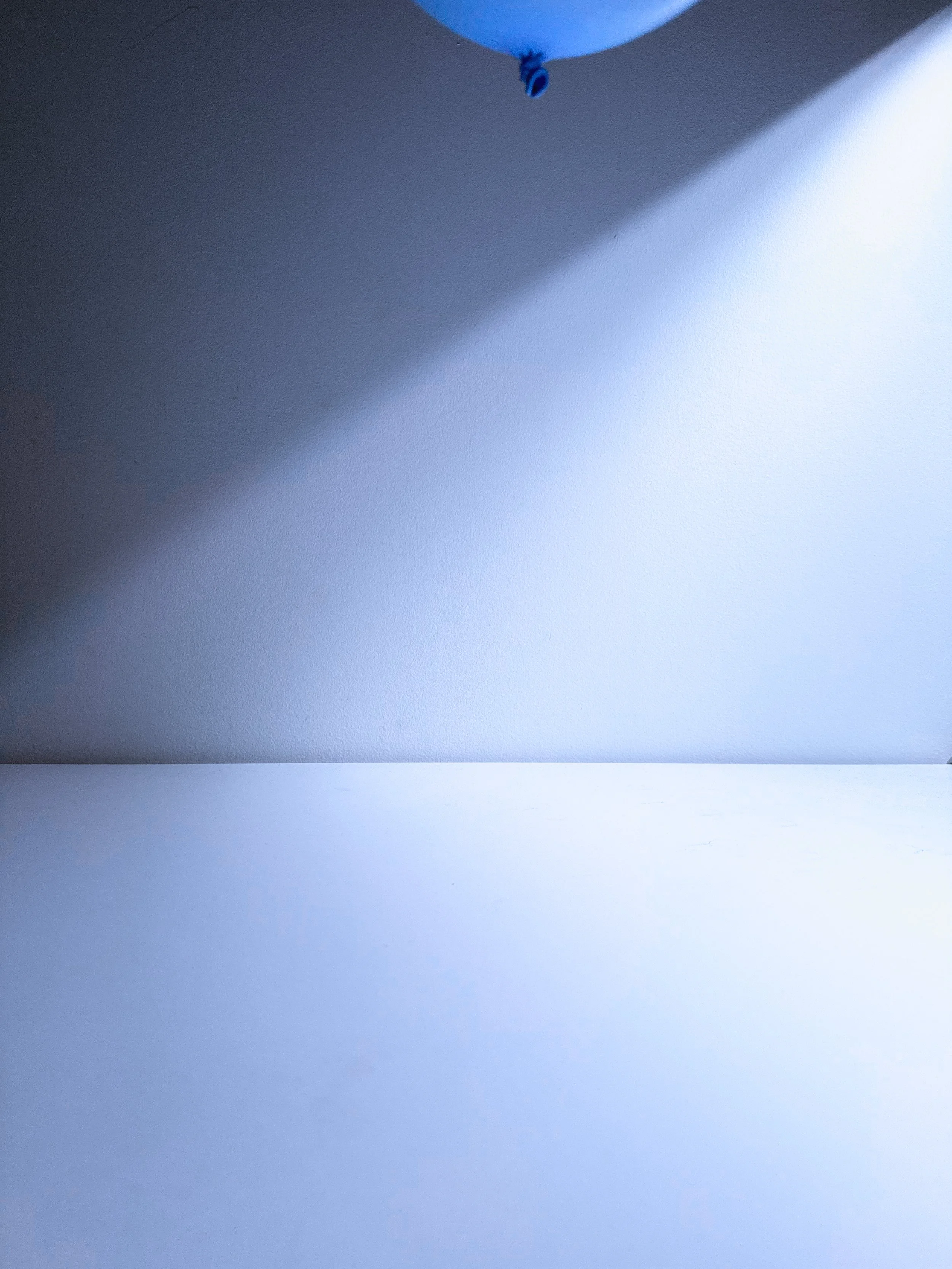 A blank wall with a small blue and black balloon tied to a ceiling hook, casting a shadow on the wall.