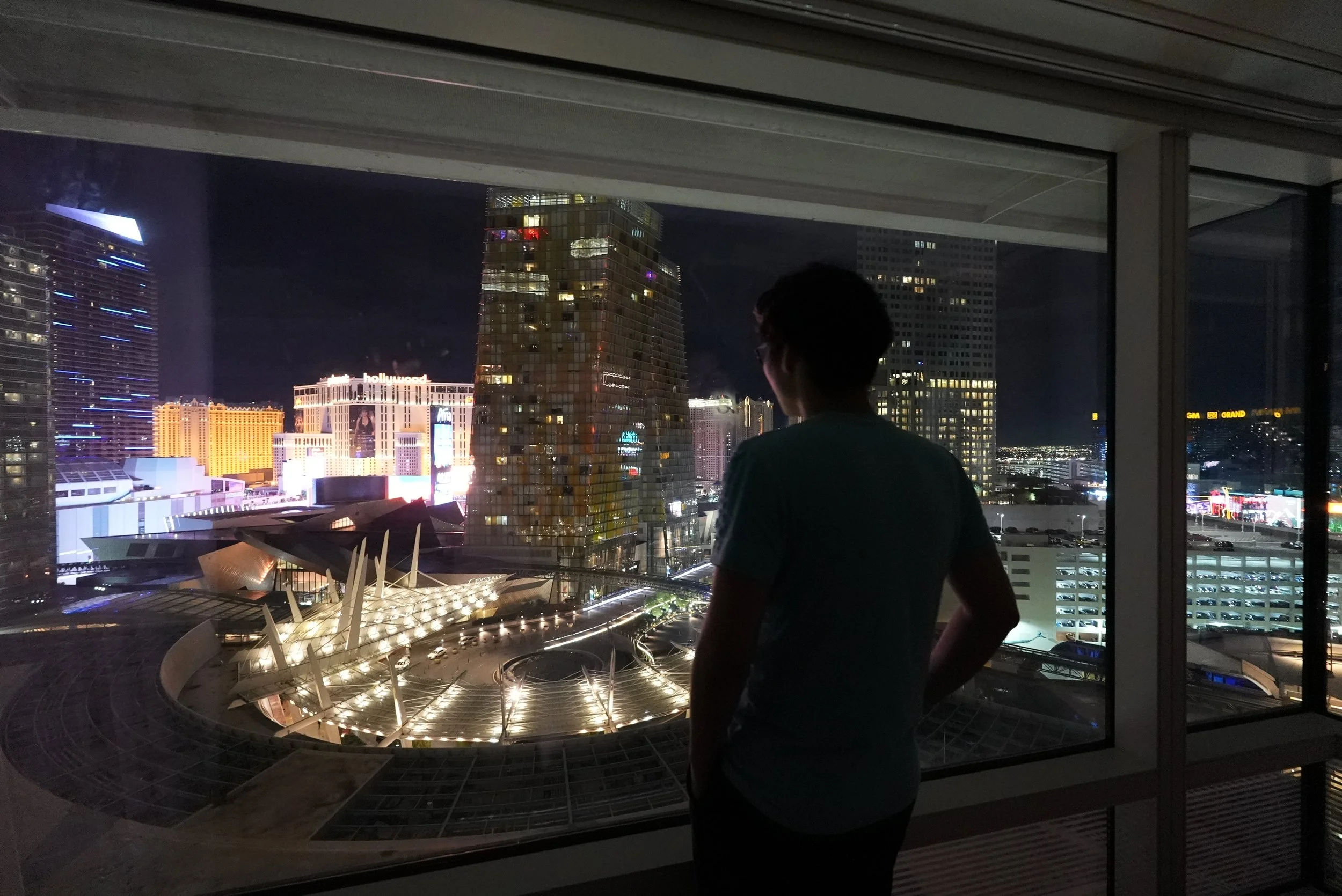 A person with dark hair and glasses stands inside a building, looking out a large window at a cityscape at night with illuminated skyscrapers and neon signs.
