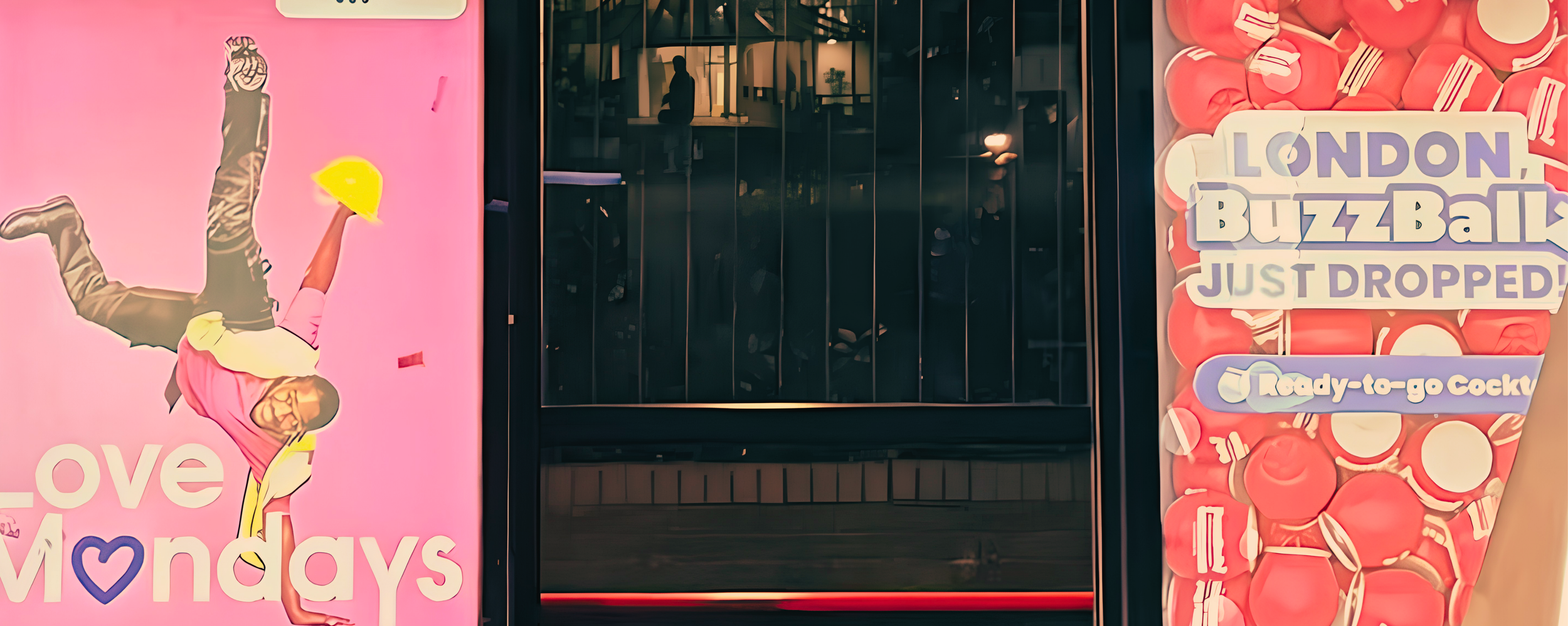 Storefront window with colorful advertisements, one featuring a person in a pink shirt holding a yellow cocktail and the other with red and white candies and the text 'London BuzzBallz Just Dropped! Ready-to-go Cocktails'
