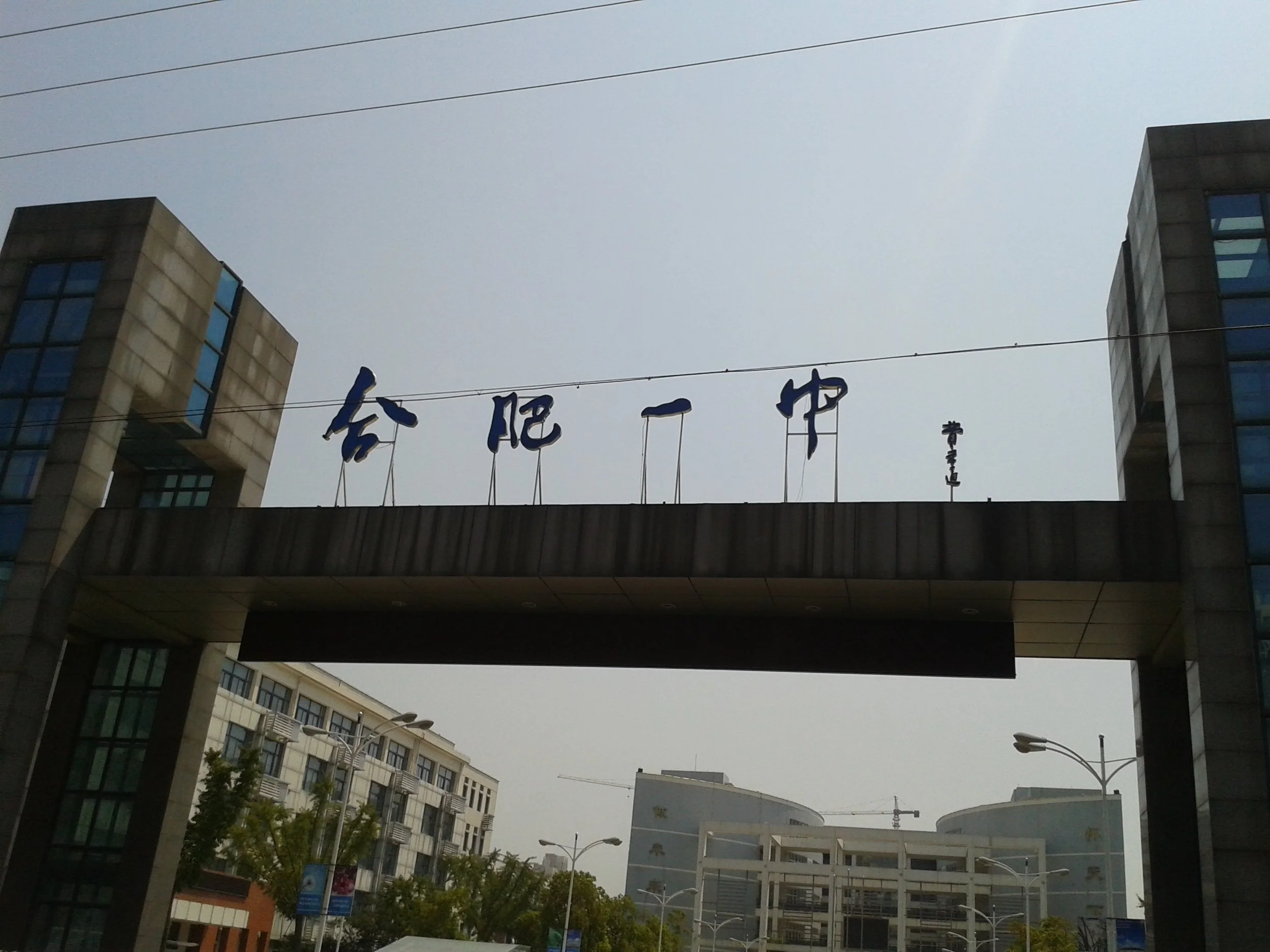 View of modern buildings with large sign with Chinese characters hanging above the street, multiple power lines, streetlights, and trees, under a cloudy sky.