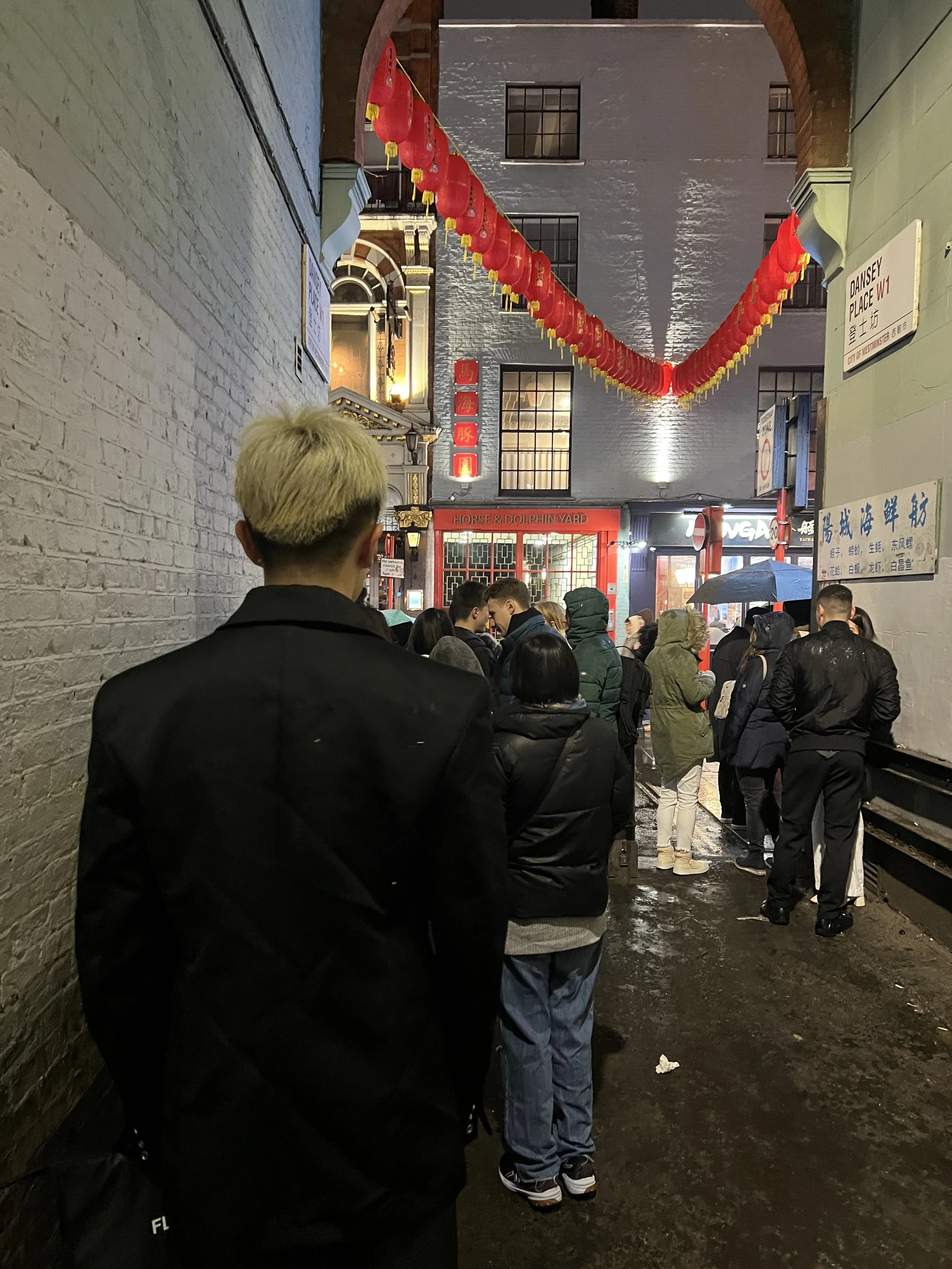 A crowded street scene at night with people standing in line outside an Asian restaurant, decorated with red lanterns hanging from the ceiling. A sign indicates the location as DANSY PLACE W1, in London.
