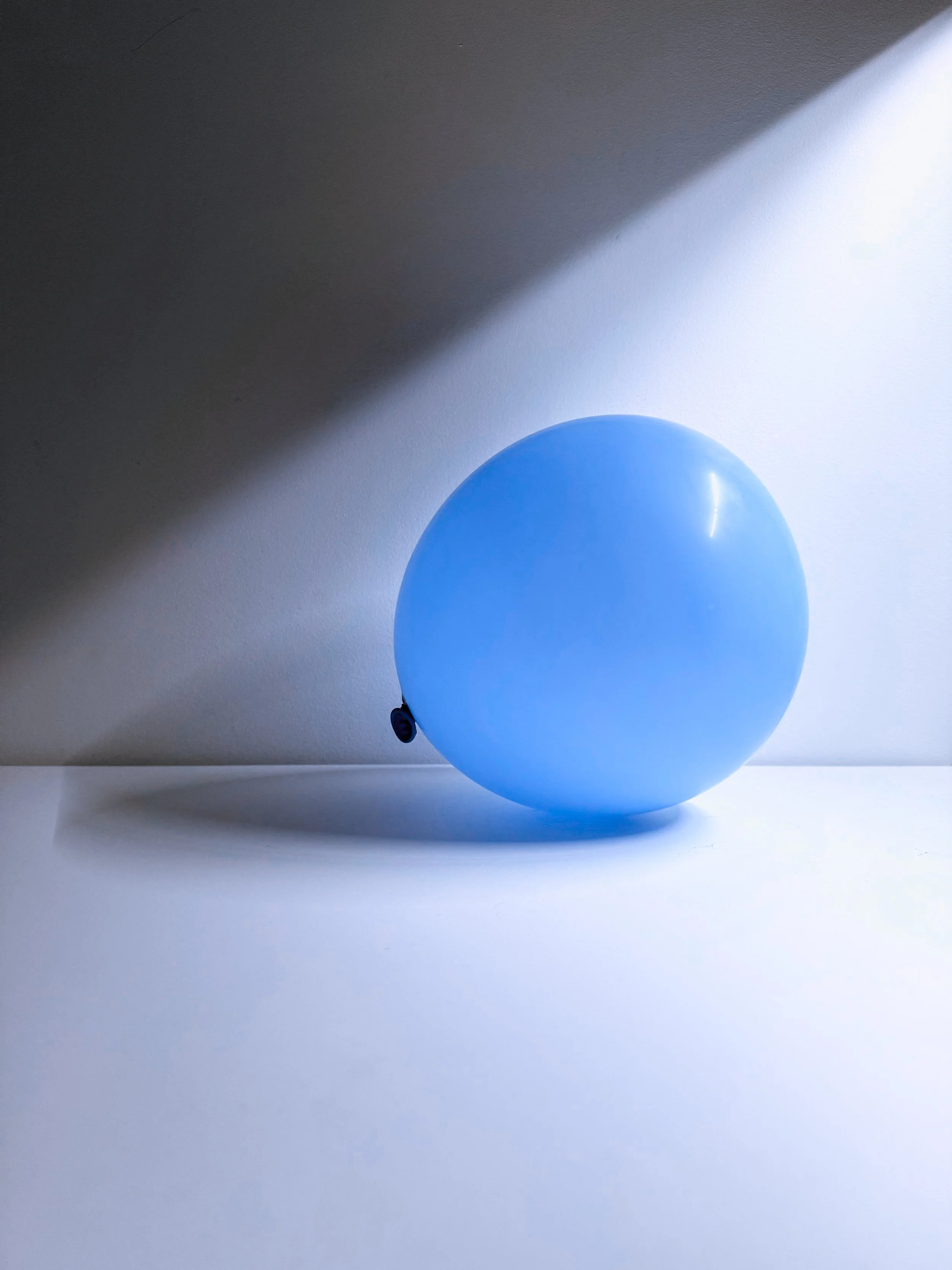 A blue balloon resting on a white surface, with light creating a bright spot on the balloon and shadows cast on the surface.