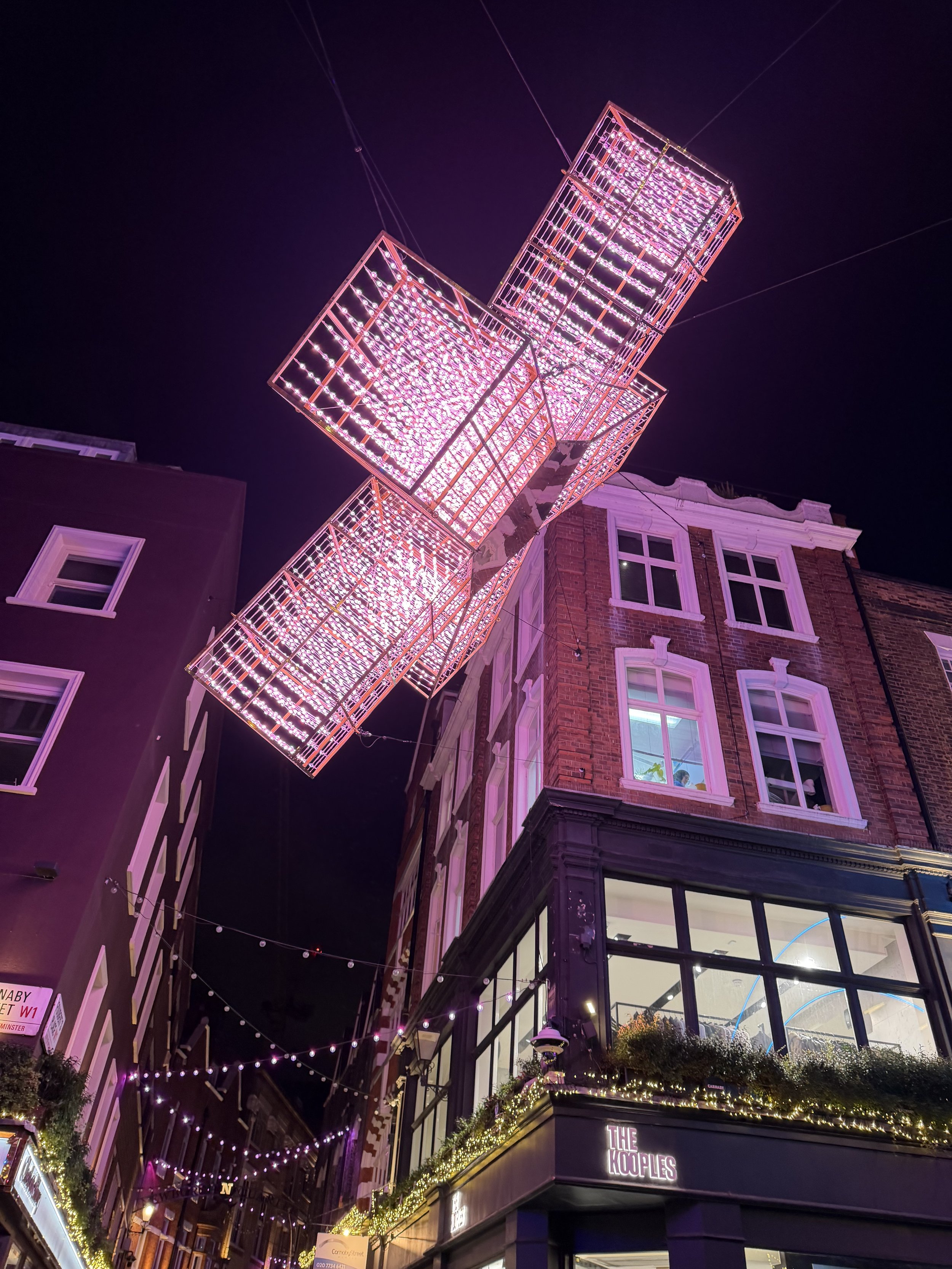 Nighttime view of a building with illuminated pink and white Christmas lights hanging from the exterior, with a narrow street below decorated with additional string lights and greenery.