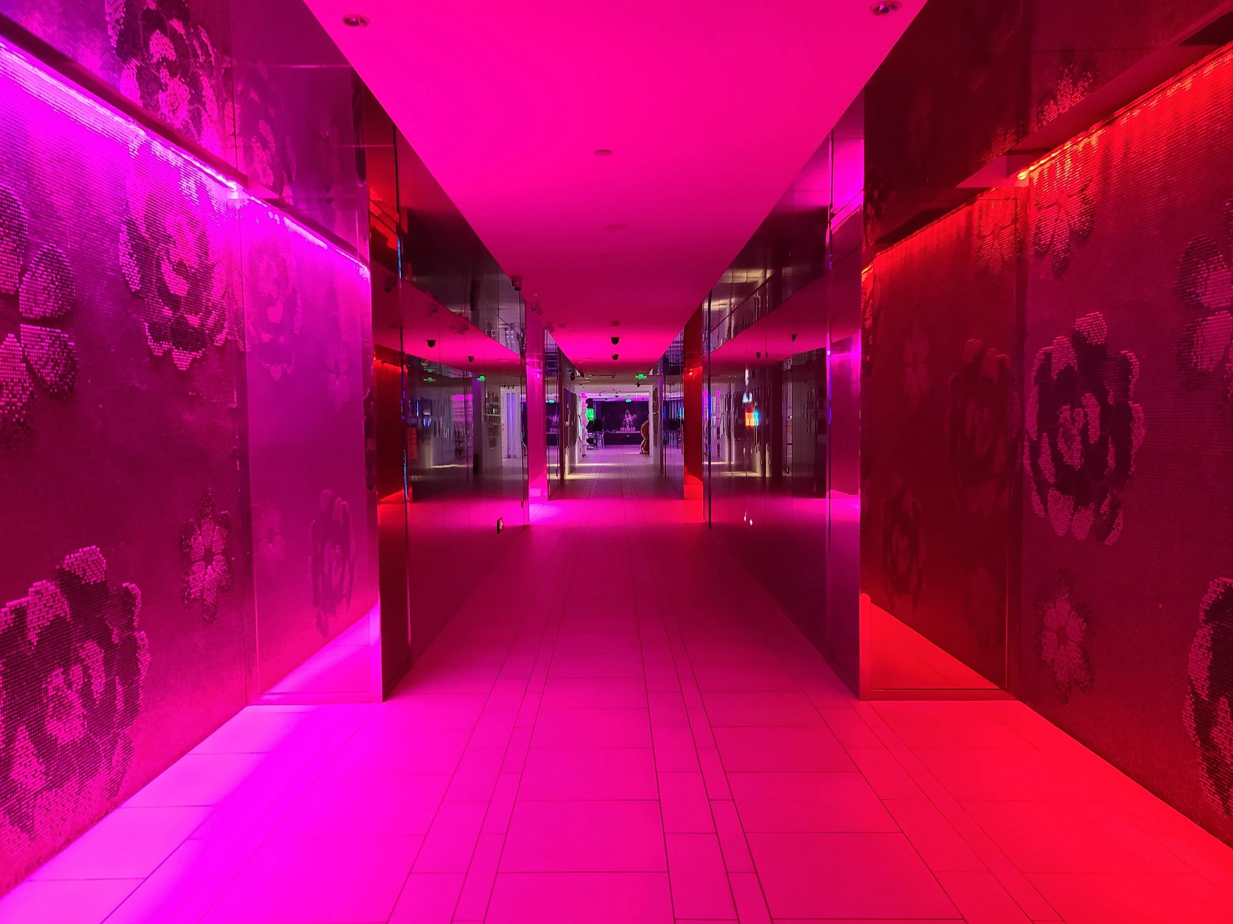 A hallway illuminated with pink and red LED lights, with reflective wall panels on both sides and a tiled floor.