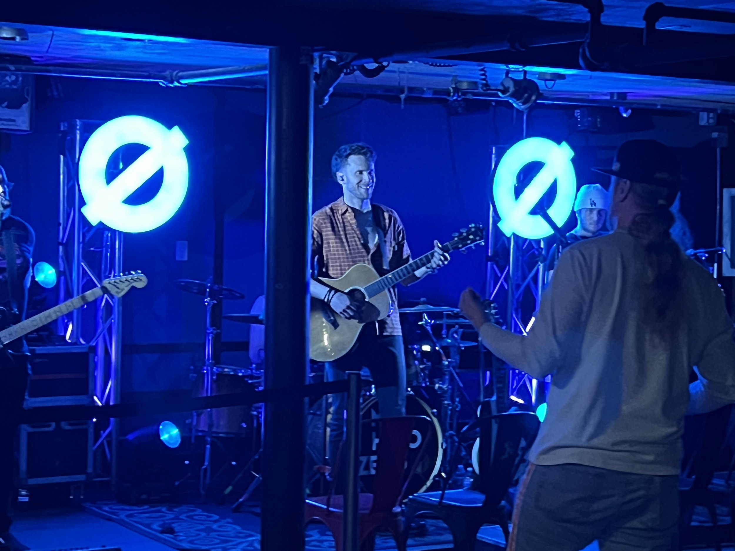 A man smiling and playing an acoustic guitar on a stage illuminated with blue lights, with neon logos behind him and people watching.