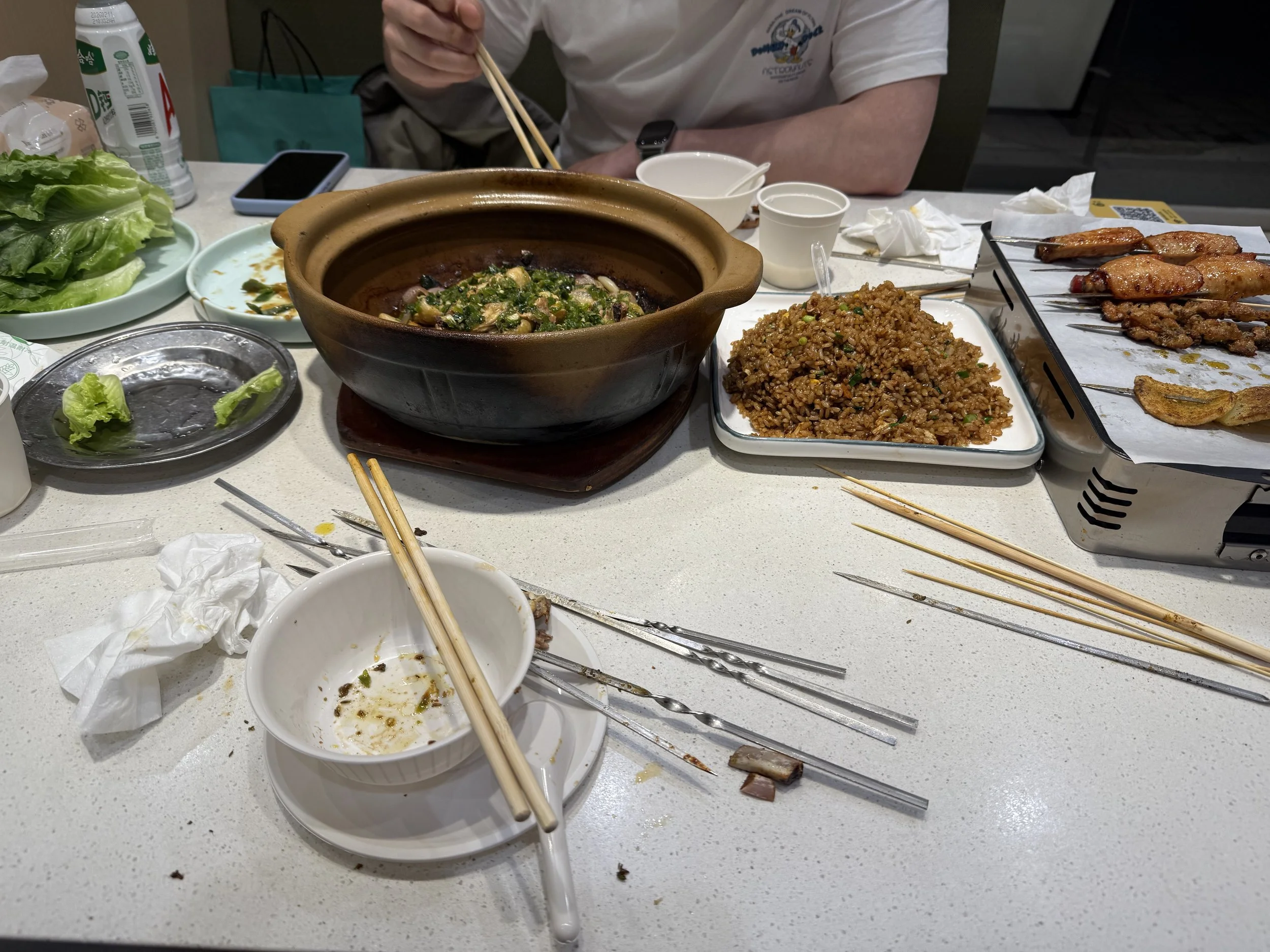 A table after a meal with empty and used dishes, chopsticks, and leftover food, including a large brown ceramic bowl with remnants of soup or stew, a white plate with a small piece of lettuce, and a small white bowl with sauce or broth. There is also