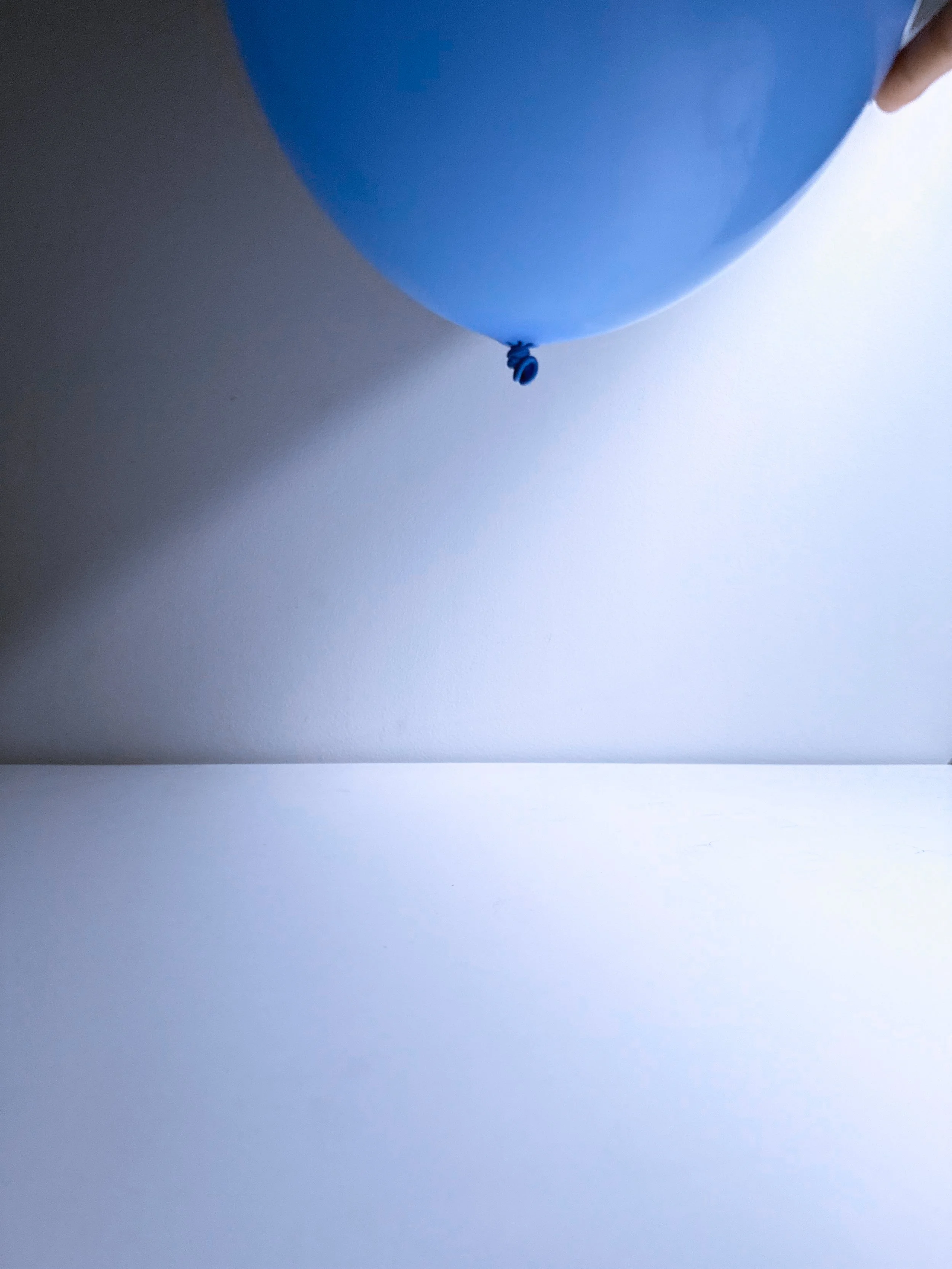 Close-up of a blue balloon tied at the bottom, with part of a hand visible on the right side, against a plain white background.