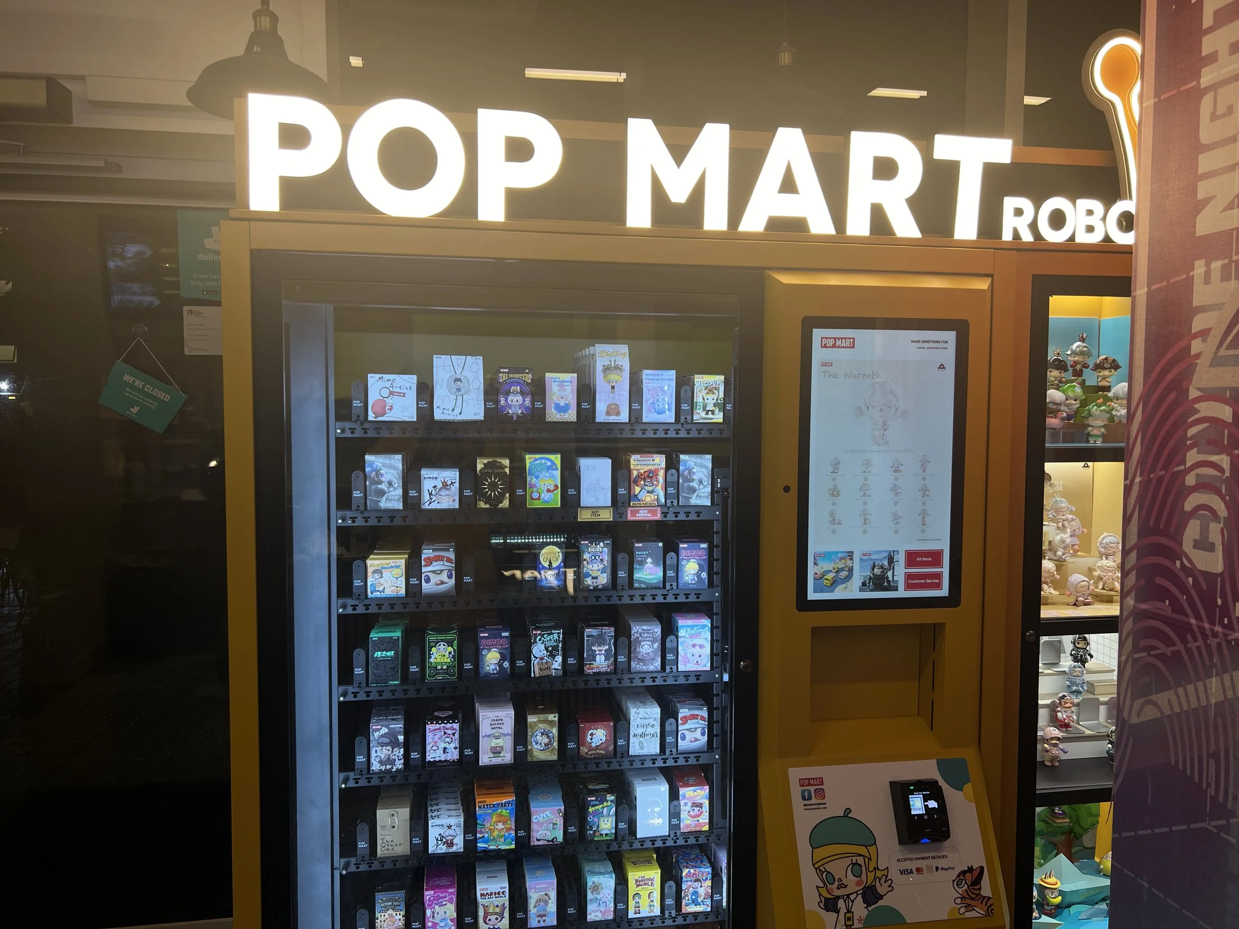 A vending machine filled with various collectible pop culture cards and figures outside a store named POP MART, with a yellow frame around it and a bright, illuminated sign.