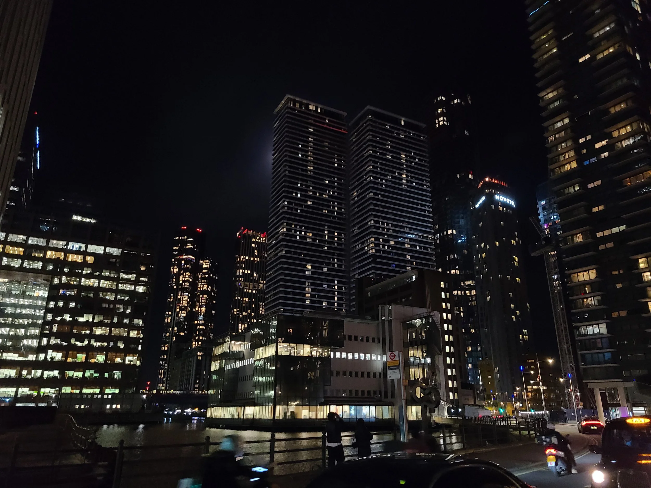 Nighttime cityscape with illuminated skyscrapers and buildings against a dark sky, with some traffic and pedestrians visible at street level.