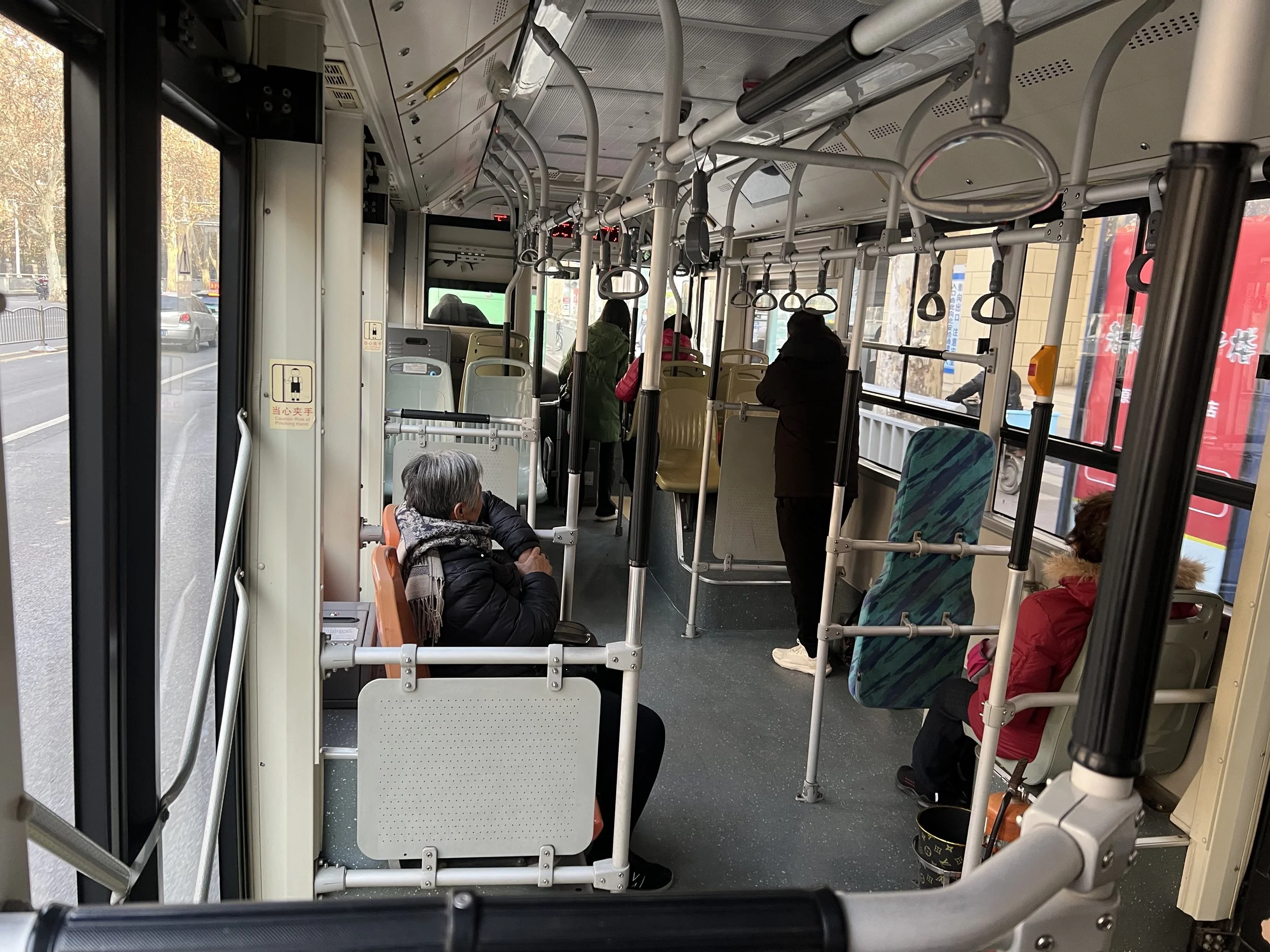 Interior of a public bus with passengers seated and standing, view through the front door, showing a road outside with trees and vehicles.