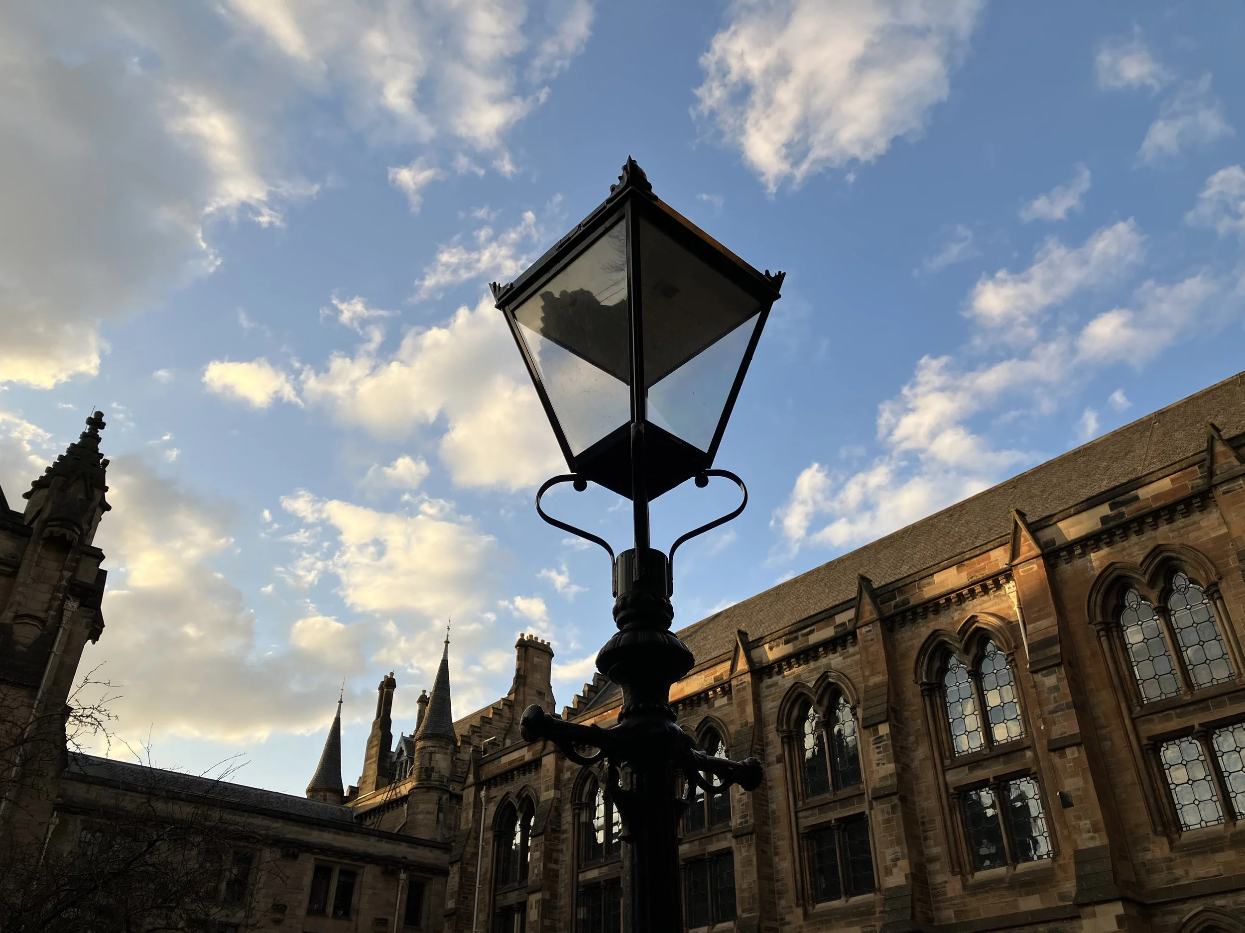A vintage black street lamp post in front of a historic building with pointed towers, set against a daytime sky with scattered clouds.