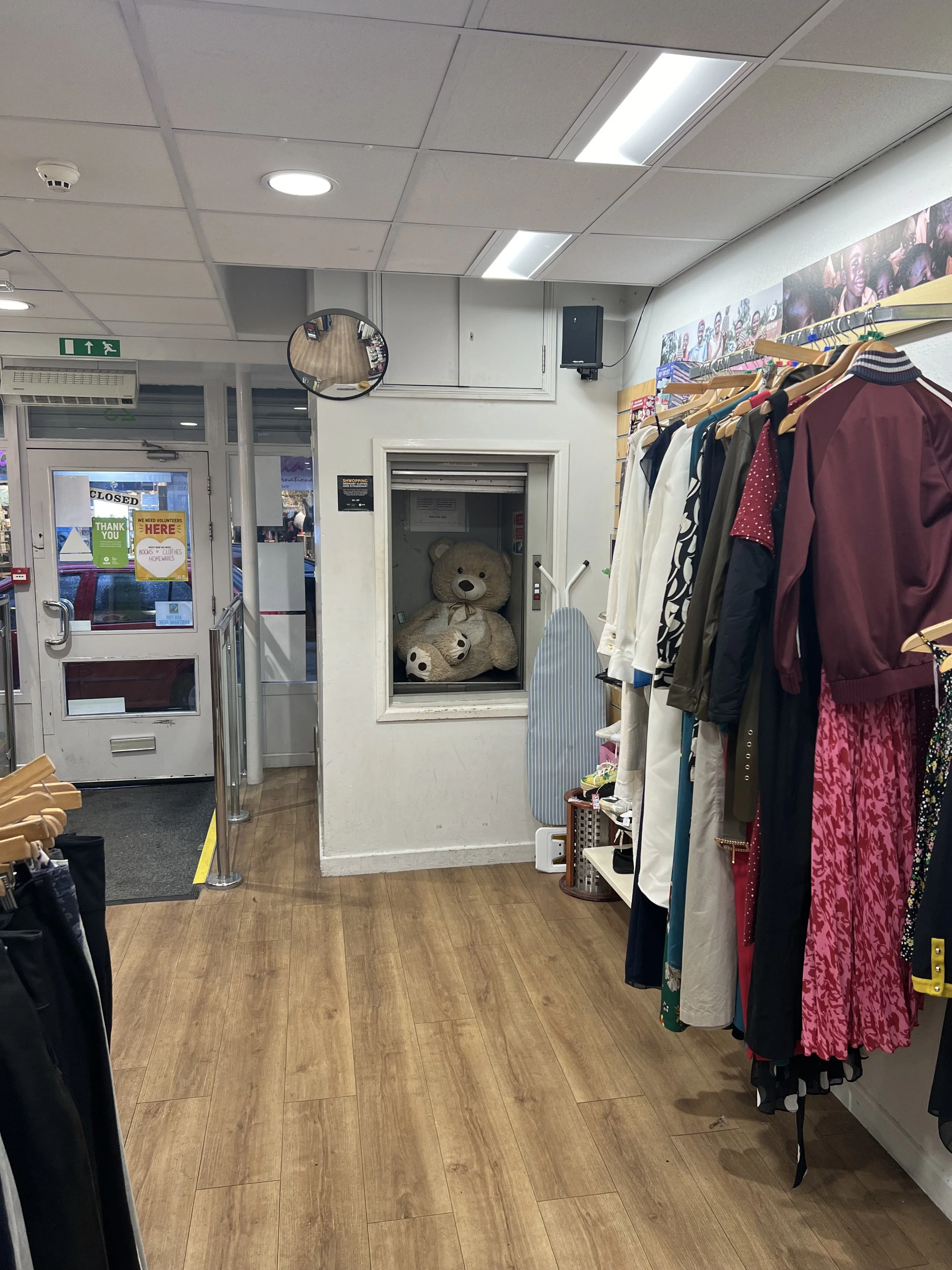 Inside a clothing store with a rack of colorful dresses and jackets on the right, a wall with photographs, a mirror, a bathroom or storage space with a teddy bear inside, and store entrance in the background with signs and a reflective surface.