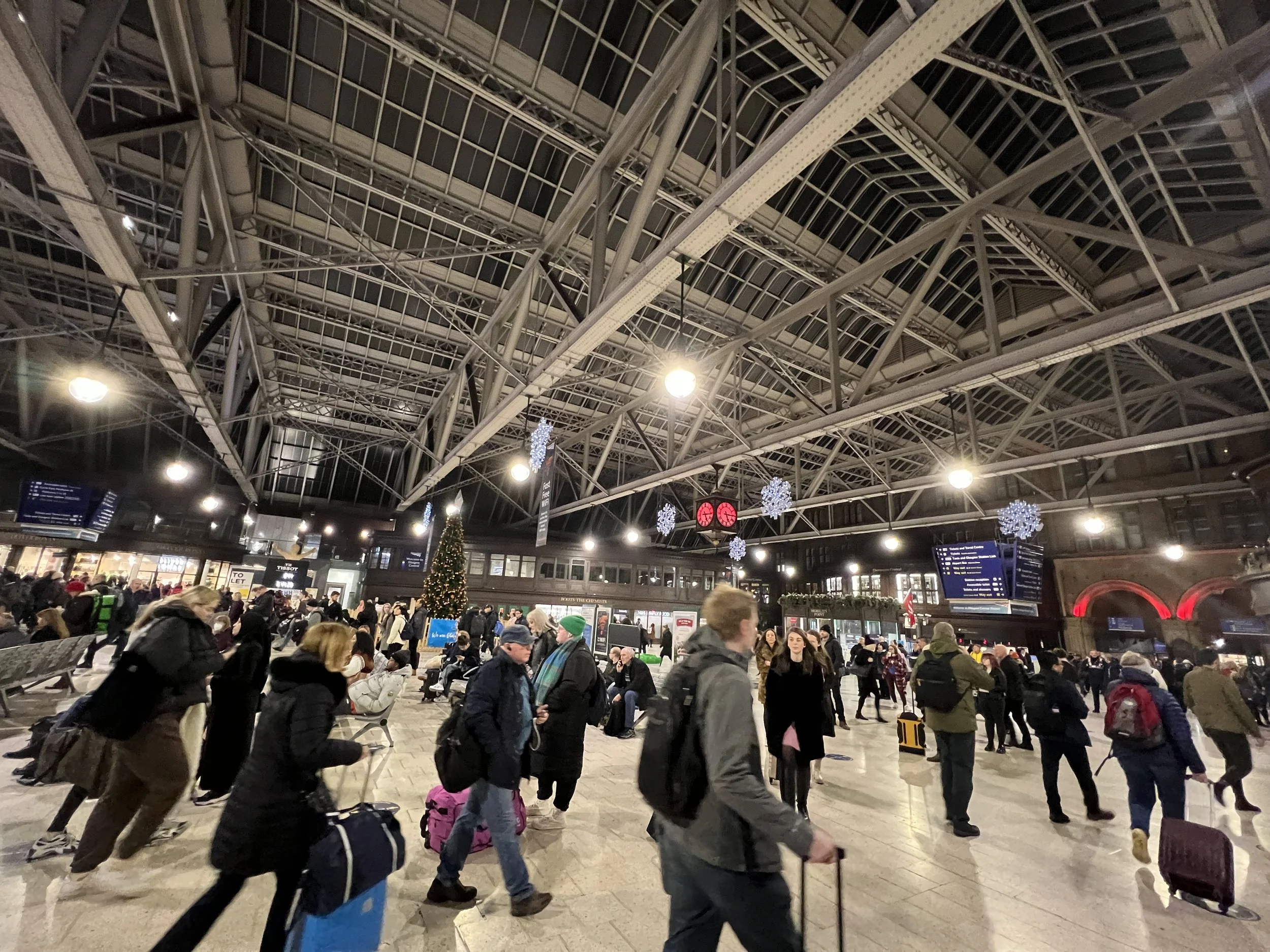 Crowded train station terminal with many people walking and waiting, festive Christmas decorations hanging from the ceiling, and a Christmas tree in the center.