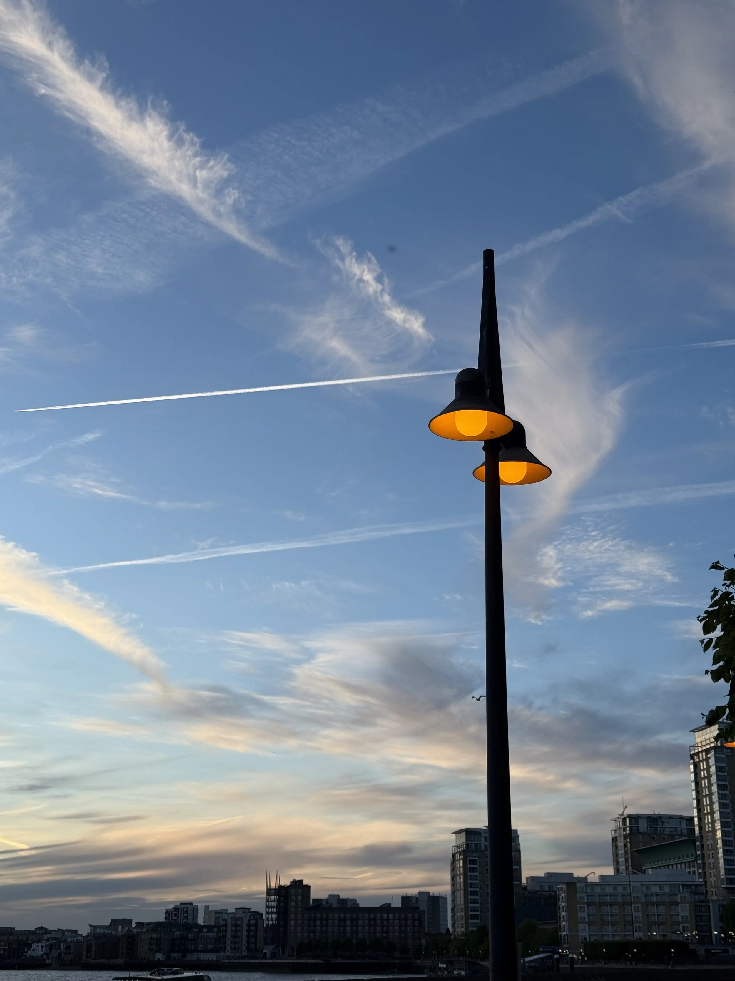 City skyline with tall buildings near a body of water, under a sky with scattered clouds and contrails, during either sunrise or sunset.
