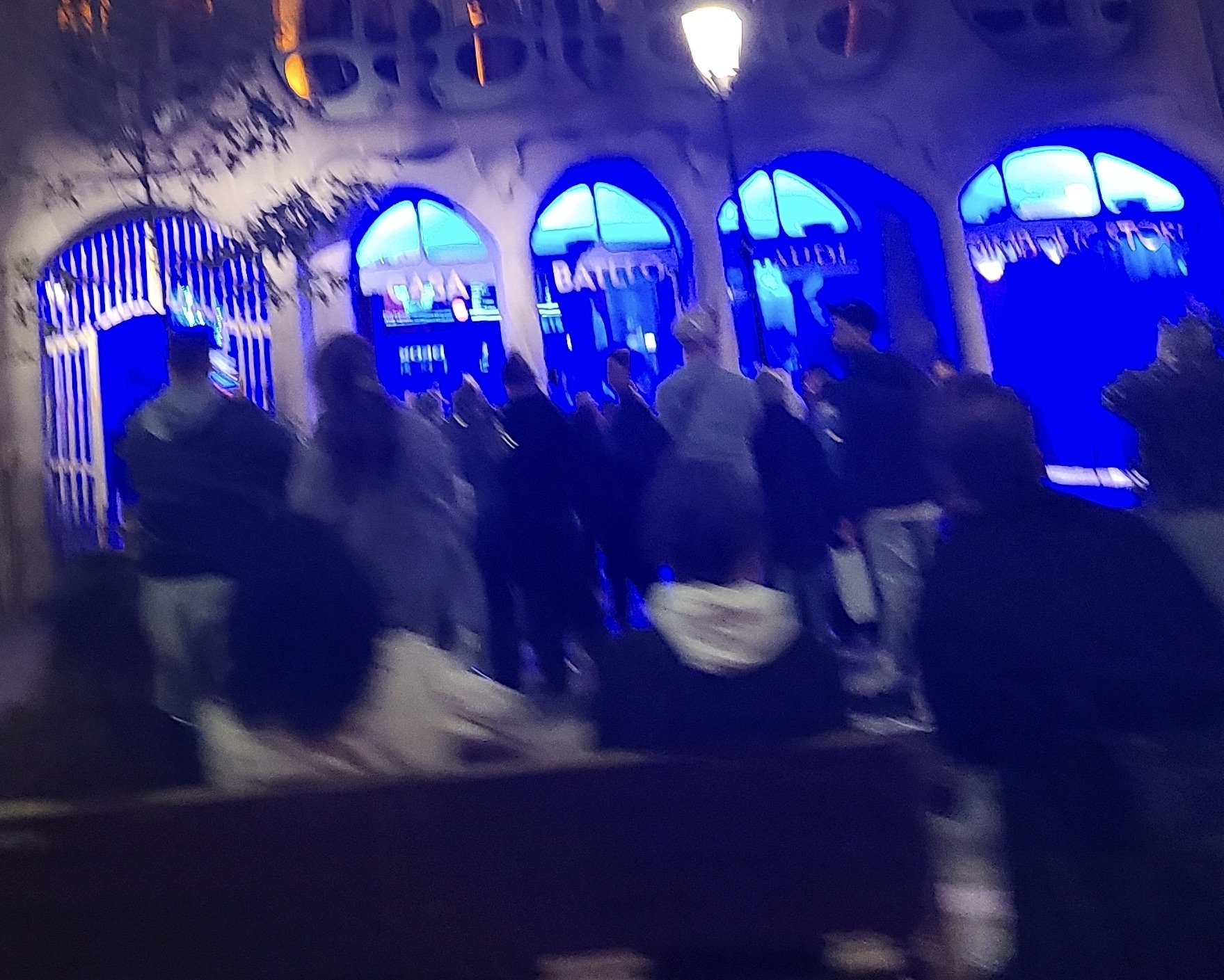 Night scene of a building with illuminated blue windows, people gathered outside, some wearing coats, under streetlights.