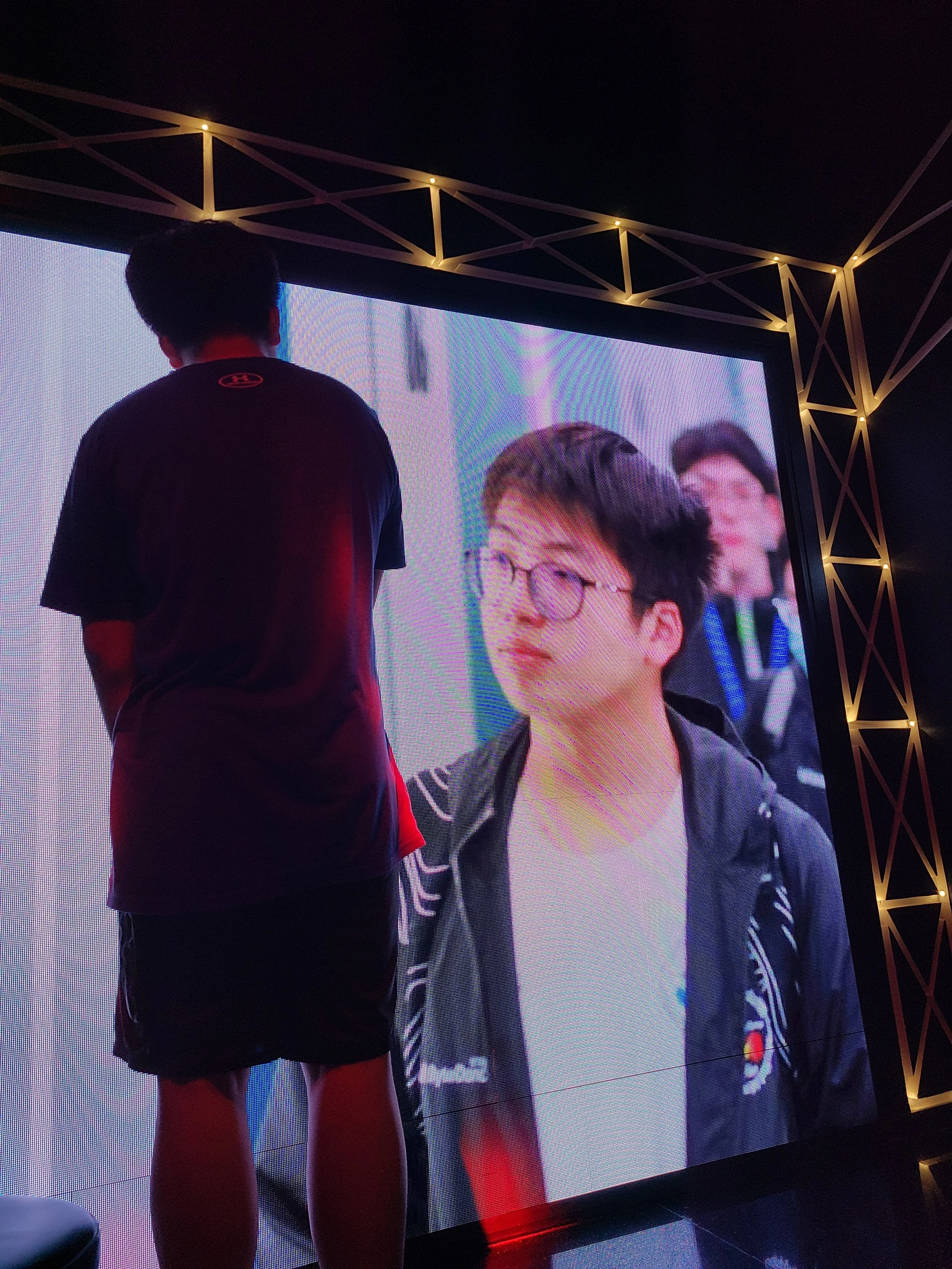A person with dark hair in a black Under Armour shirt and shorts standing in front of a large digital screen showing a man with glasses and dark hair, possibly in a professional setting.