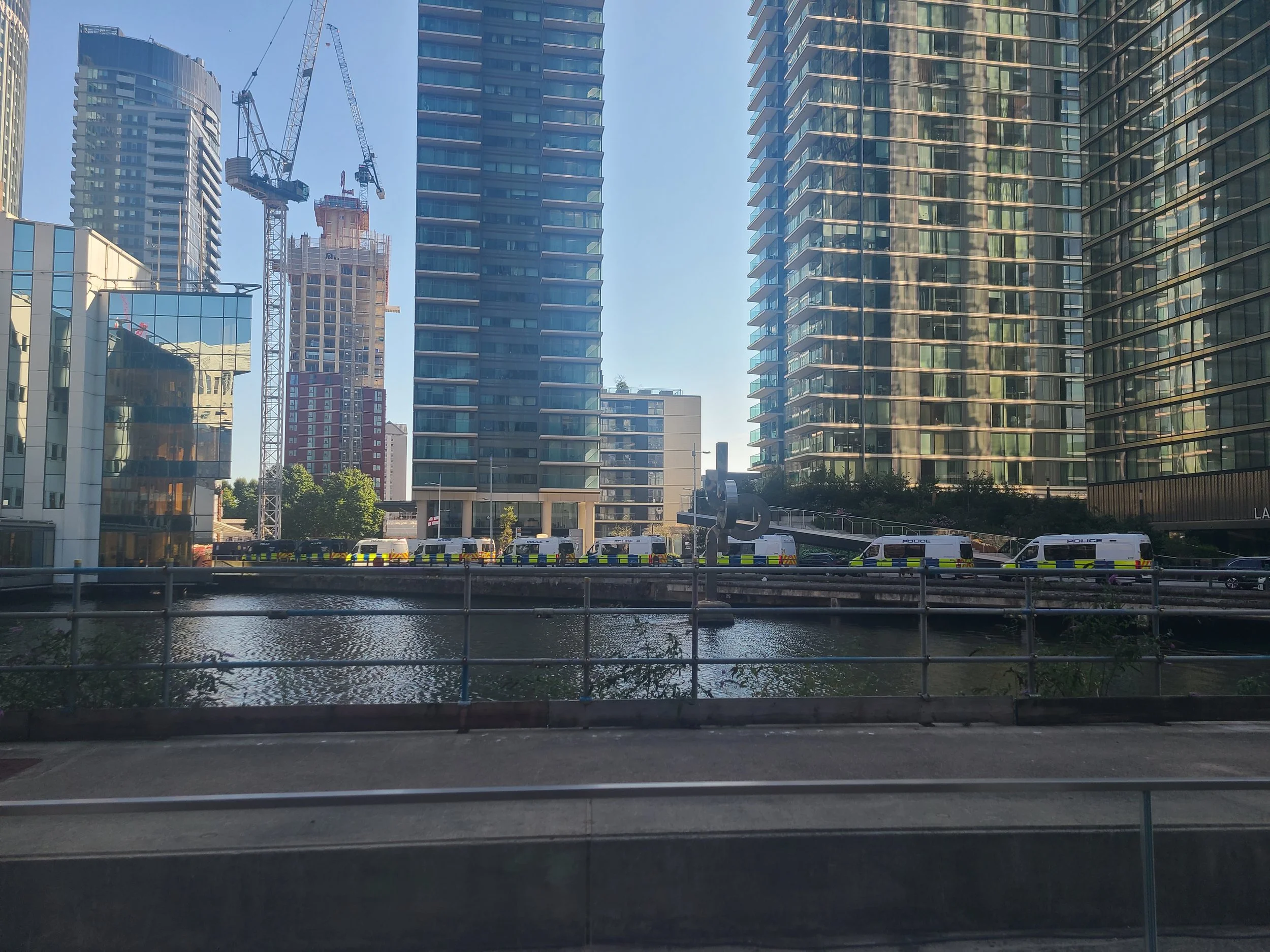 Cityscape with several modern high-rise buildings and construction cranes, viewed across a river, with police vehicles parked along the street in the foreground.