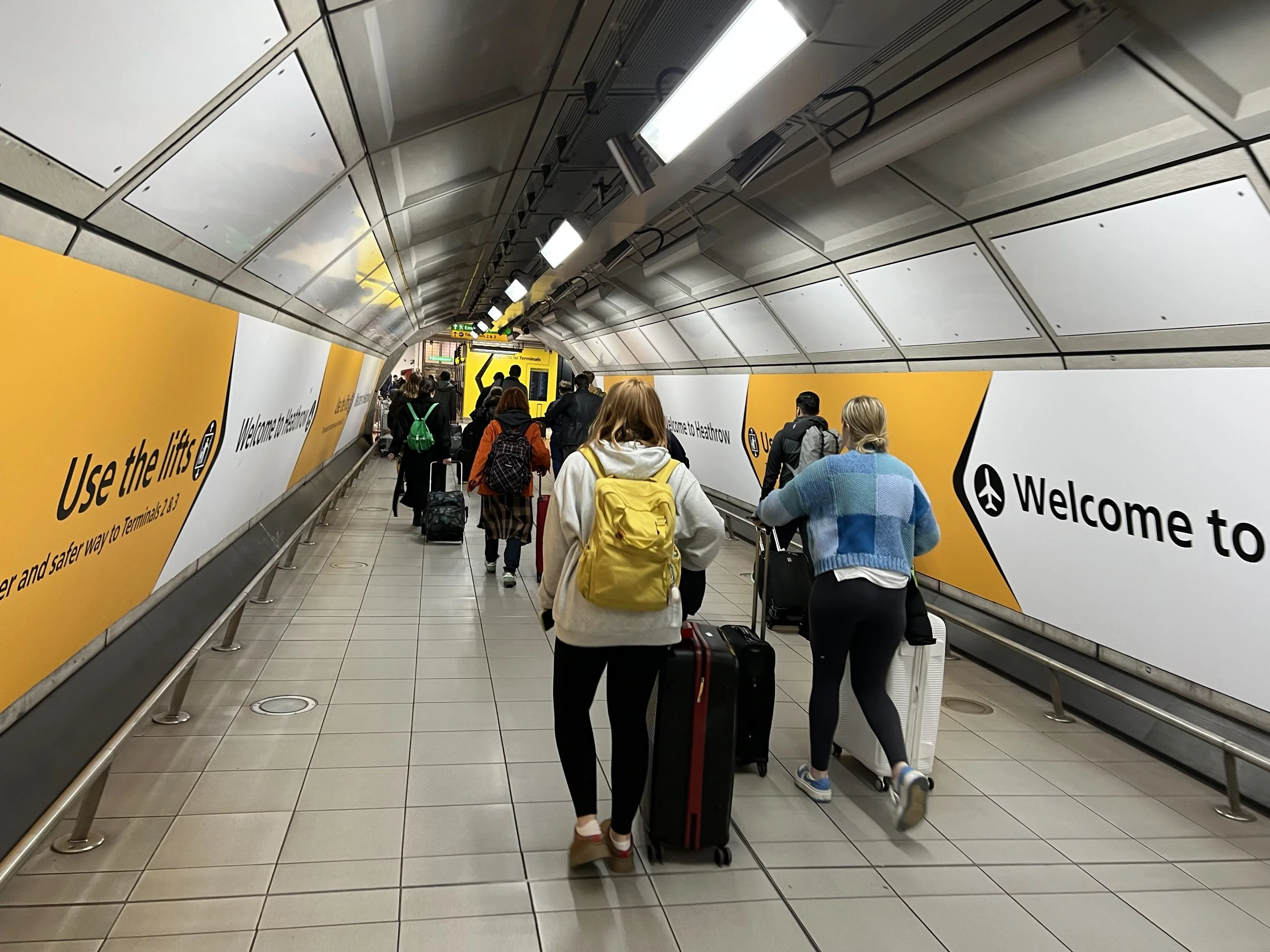 People walking in an airport terminal tunnel with luggage, yellow signs and advertisements along the sides.